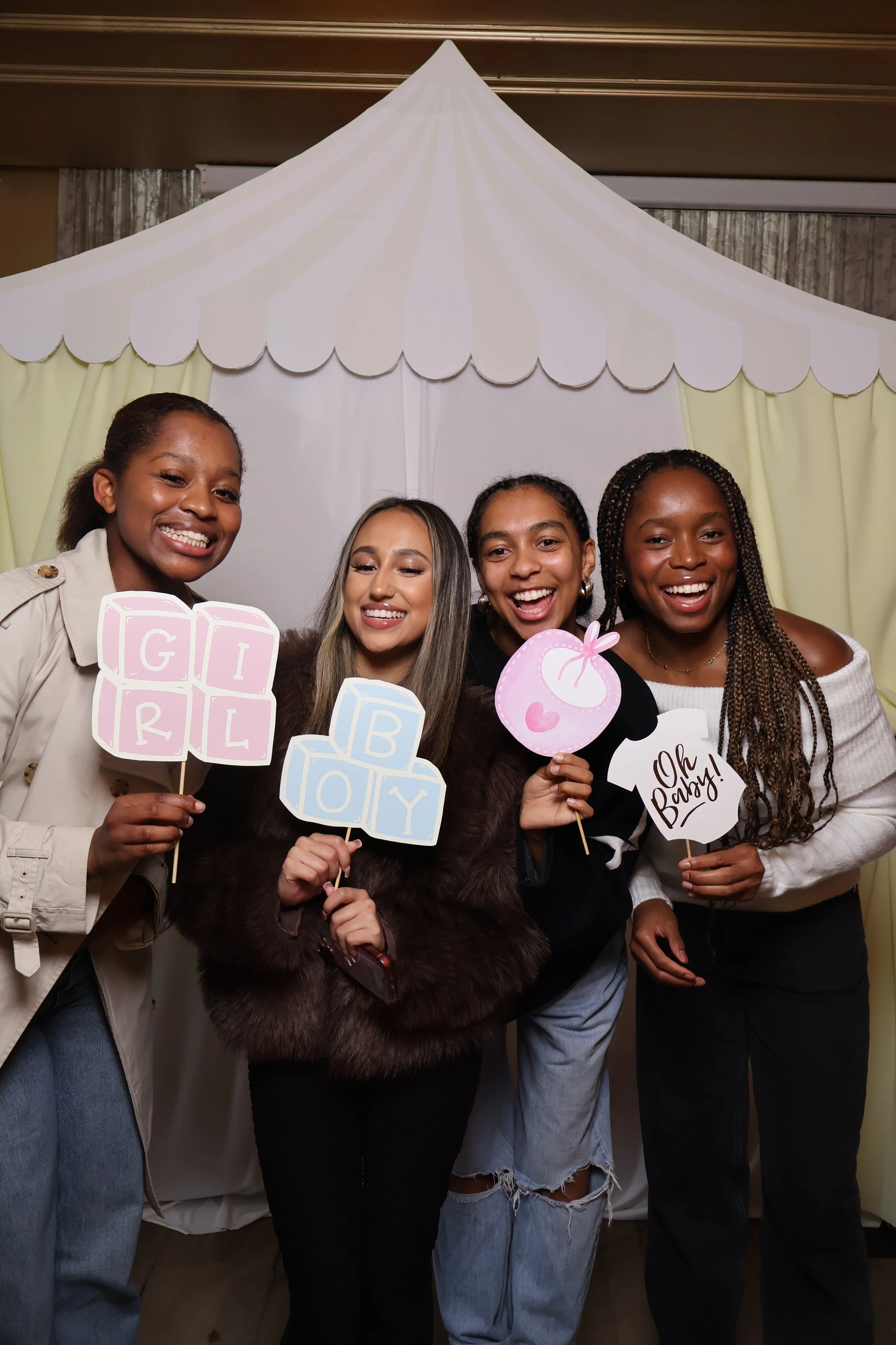 Four women smiling and holding baby shower signs, standing in front of a white tent backdrop.