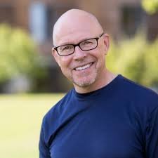A smiling man with glasses and a shaved head outdoors on a sunny day.