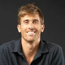 A young man with light brown hair and a beard, smiling, wearing a dark button-up shirt against a black background.