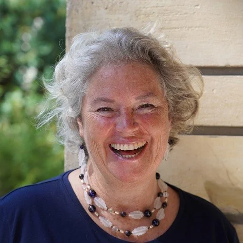 A smiling older woman with gray hair, wearing a navy blue top and a multi-strand pearl necklace, standing outdoors next to a light-colored building.