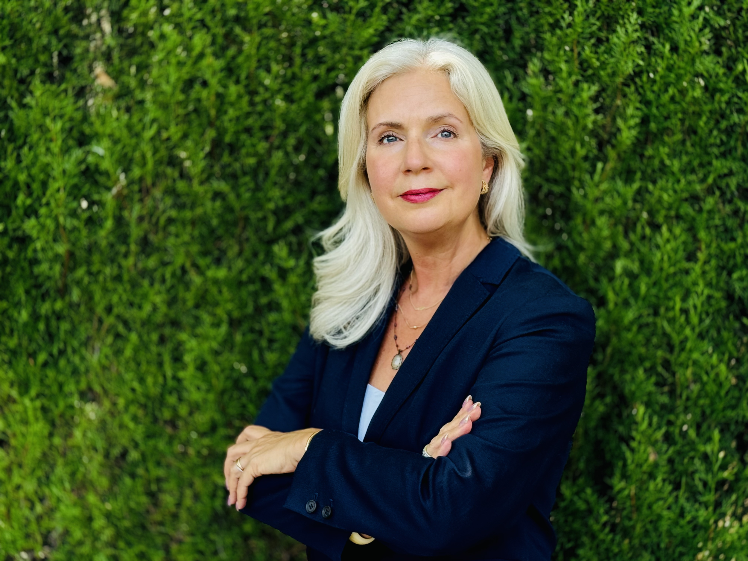 A professional woman with long blonde hair wearing a navy blazer, standing with arms crossed in front of a green hedge, looking confidently at the camera.