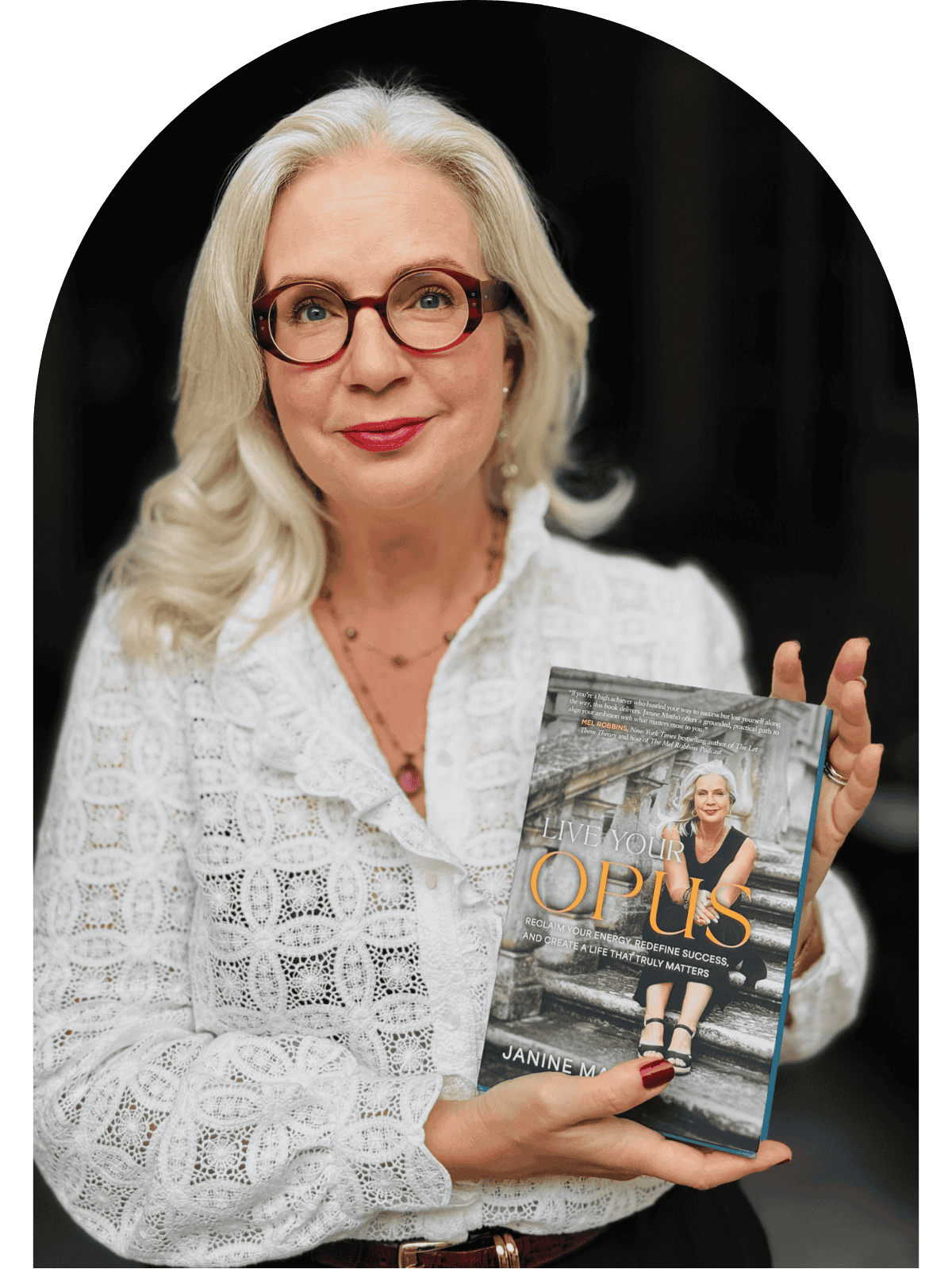 A woman with long white hair, glasses, and red lipstick holding a book titled 'Live Your Opus' by Janine M. is standing in front of a black background.