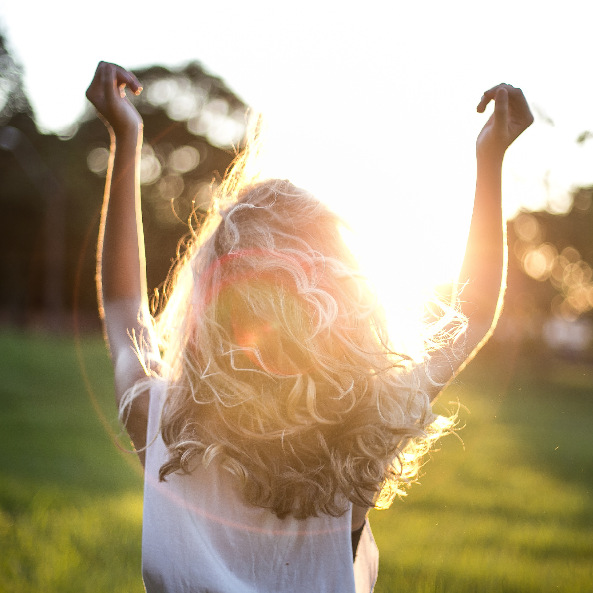 A person with curly blonde hair standing outdoors during sunset with arms raised, back to the camera, sunlight illuminating their hair.