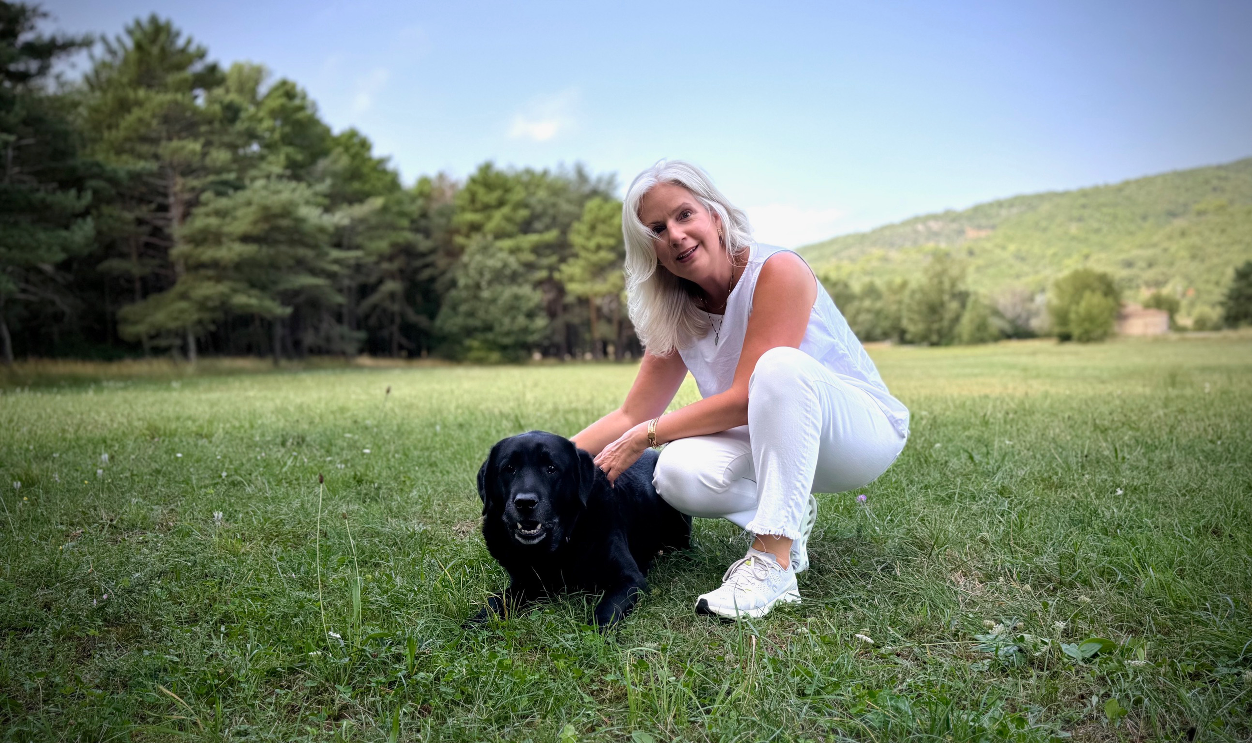 A woman with white hair and a white outfit kneels on the grass next to a black Labrador dog in a green field with trees and hills in the background.