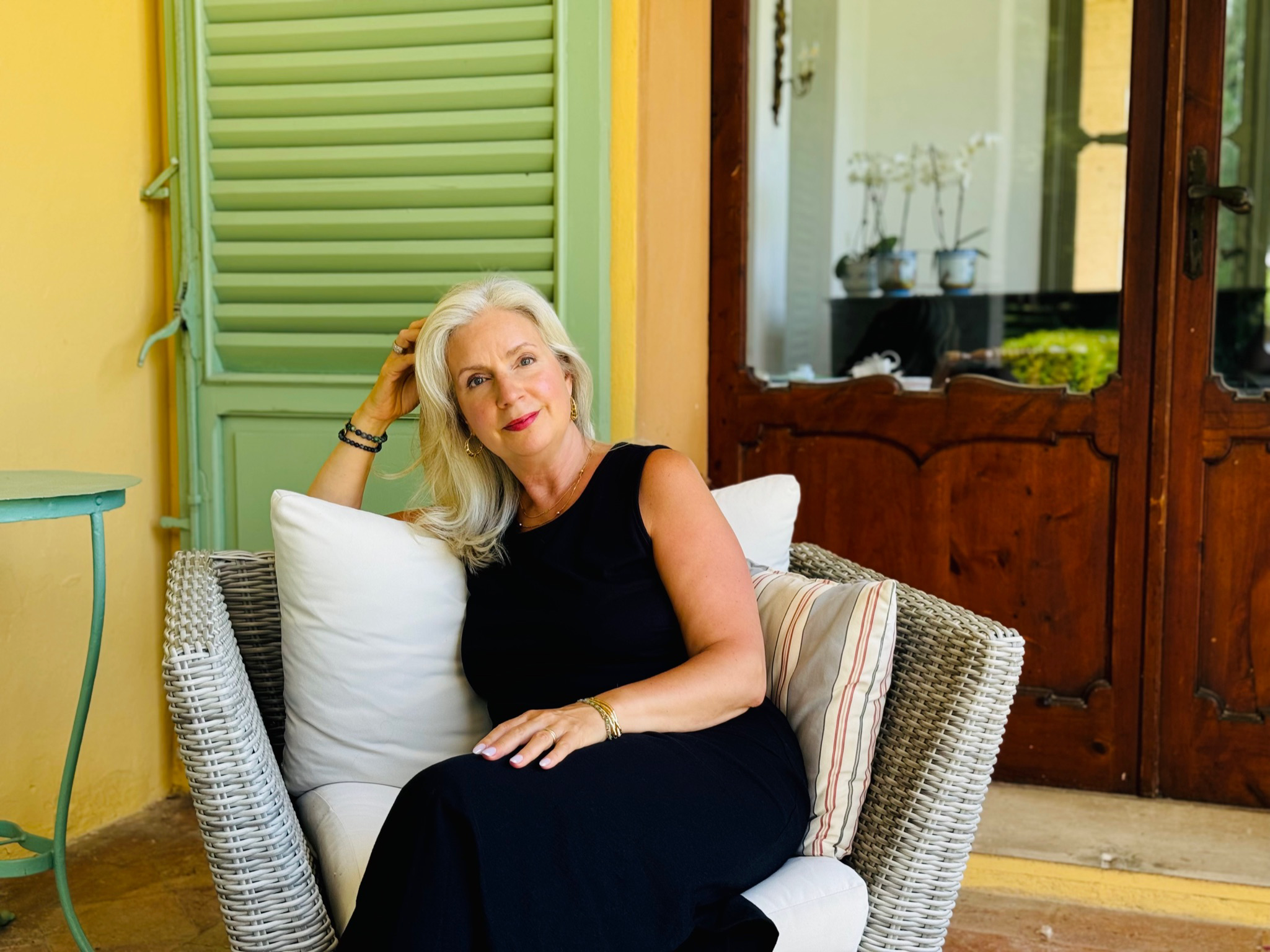An older woman with long gray hair lounging on a wicker chair, wearing a black sleeveless dress and jewelry, in a brightly colored room with yellow walls, a green shutter, and wooden cabinet doors.
