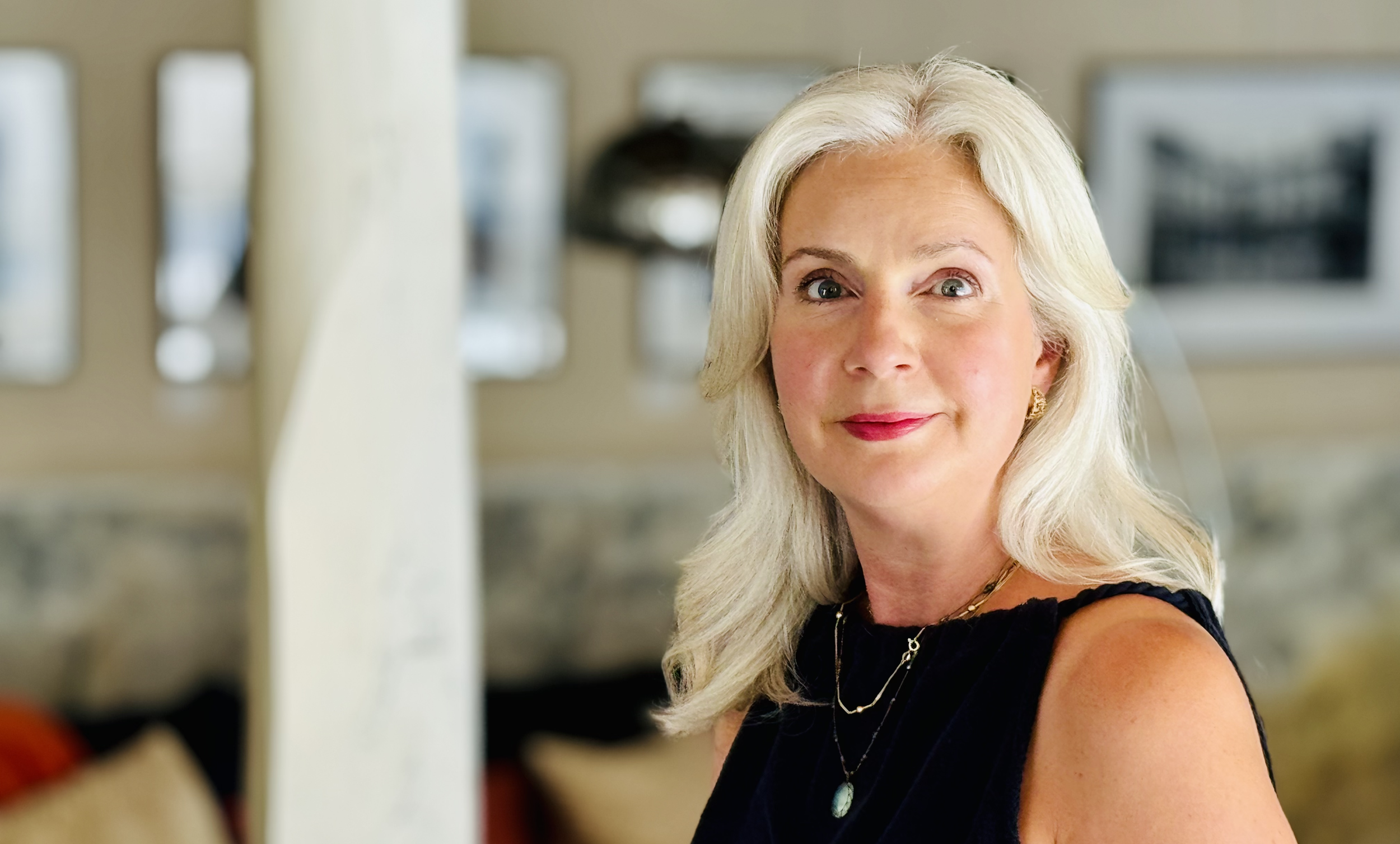 Portrait of an elegant older woman with blonde hair, wearing a black dress and layered necklaces, smiling softly in a cozy indoor setting.