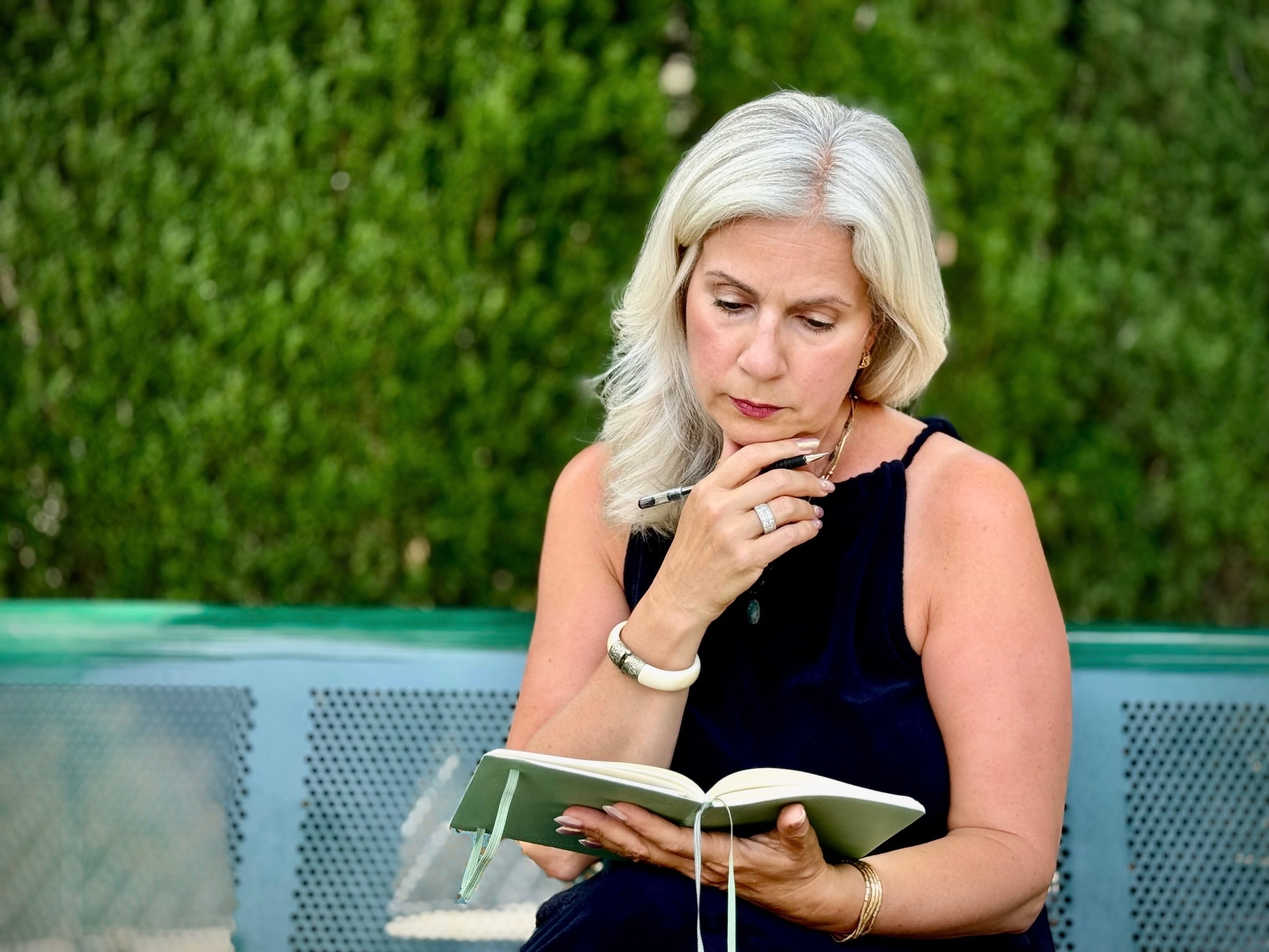 A woman with blonde hair sitting on a bench outdoors, reading a notebook, with a contemplative expression.