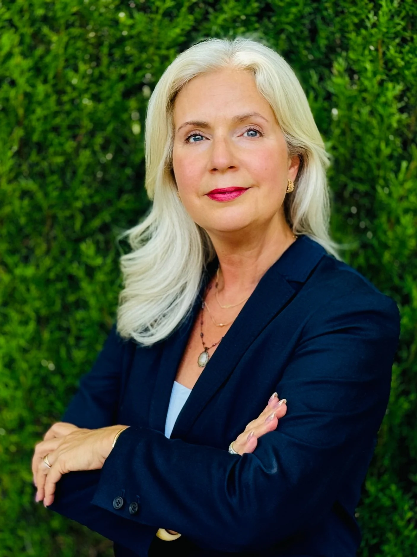 A middle-aged woman with long, wavy gray hair, wearing a dark blazer and jewelry, standing outdoors with a green hedge in the background.