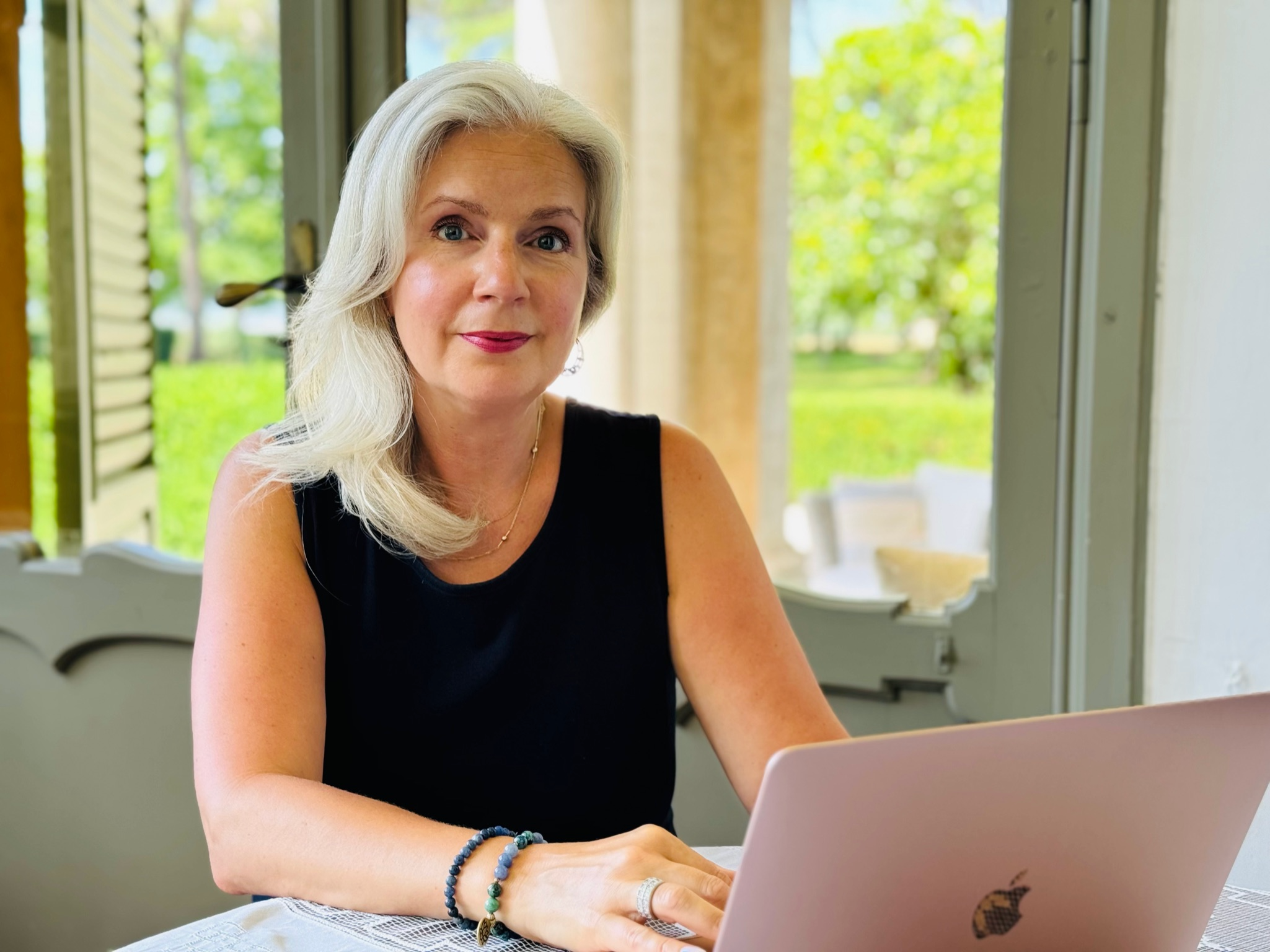 A woman with silver hair working on a pink laptop at a table indoors with open windows showing greenery outside.