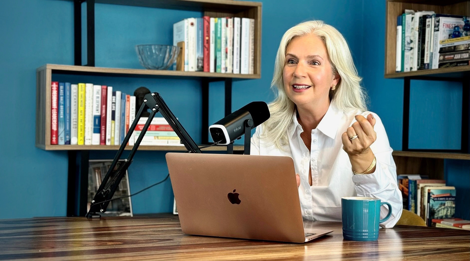 A woman with blonde hair sitting at a wooden desk in front of a laptop and a microphone, smiling and gesturing with her right hand, with bookshelves in the background.