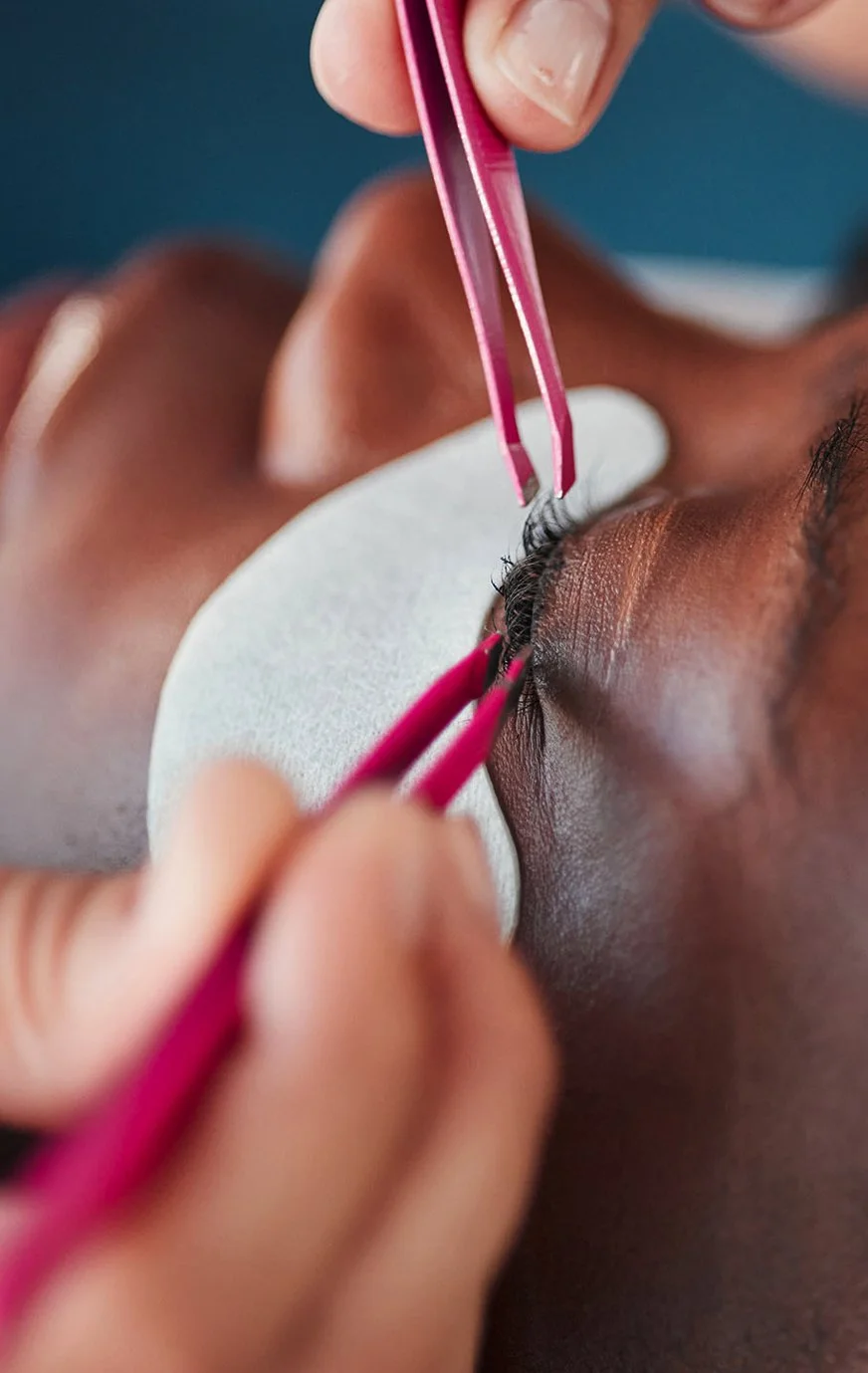 Close-up of a person receiving eyelash extension application, with beauty technician using pink tweezers to attach synthetic lashes to natural lashes, and a white pad placed under the eye.