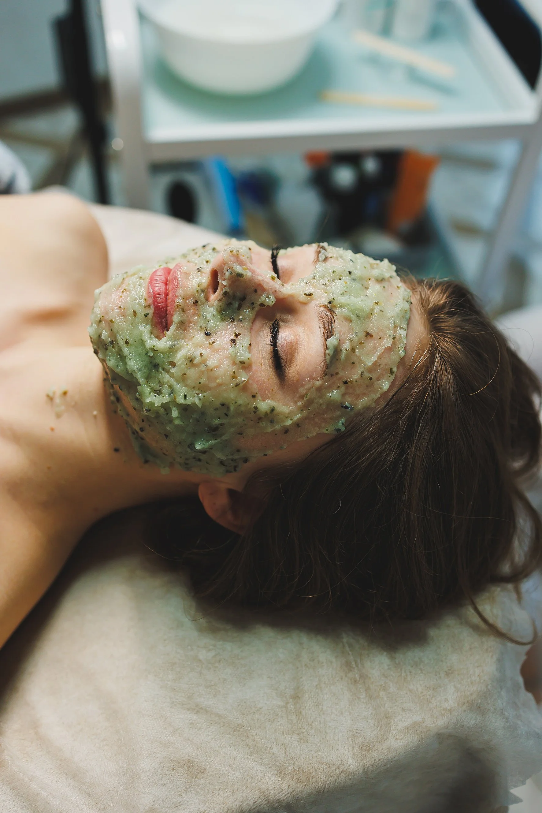 A woman lying on a hospital bed with a green clay mask on her face.
