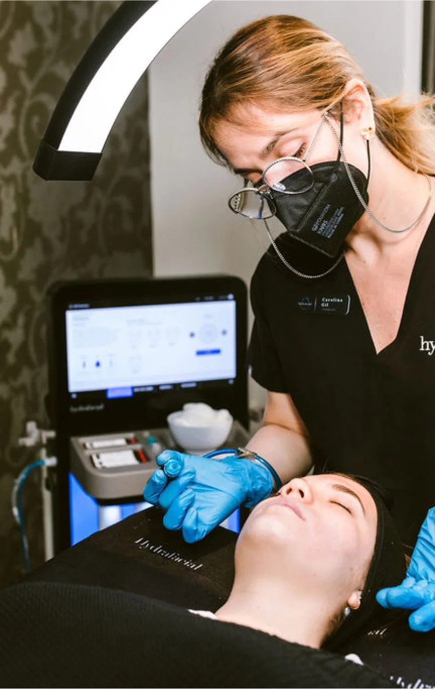 A woman wearing a black face mask and blue gloves performs a facial treatment on a woman lying down with her eyes closed in a skincare clinic.