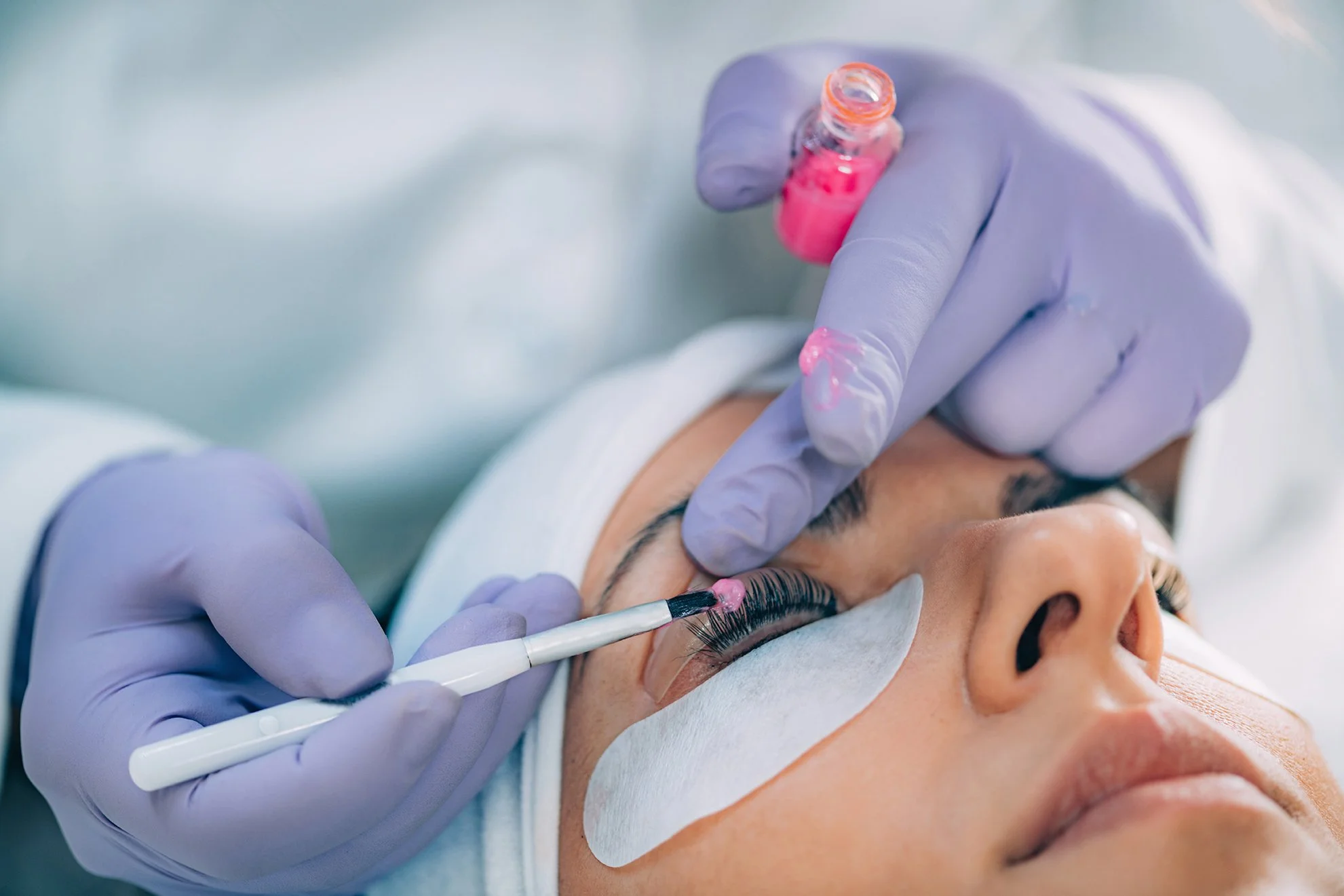 A close-up of a woman receiving eyelash extension treatment, with a technician applying pink adhesive to her upper lashes using a small brush, while wearing purple gloves.