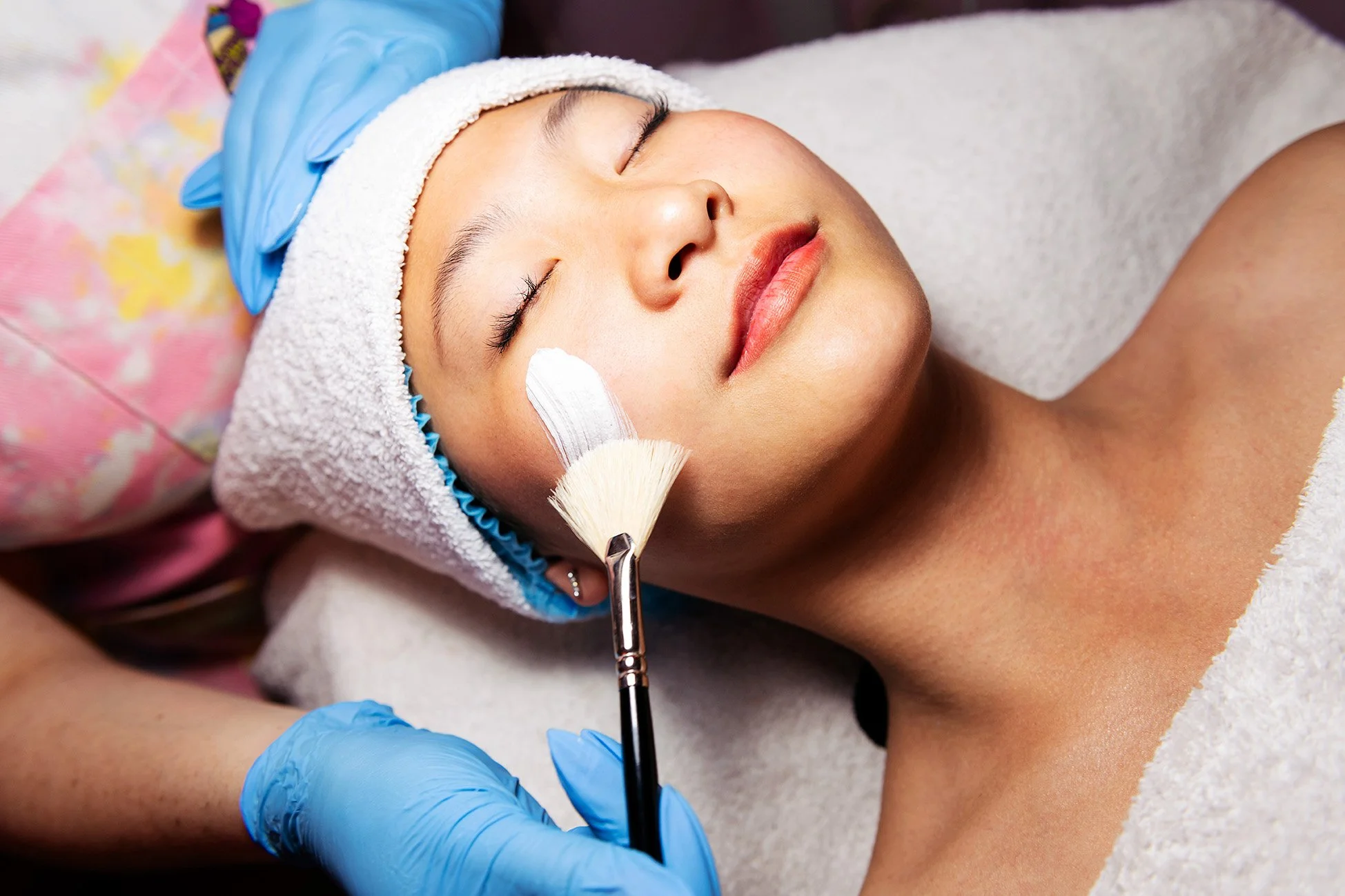 A woman lying on her back with her eyes closed, receiving a facial treatment at a spa or skincare clinic. She is wearing a white towel around her head and a gray towel on her shoulder. A skincare specialist wearing blue gloves applies a cream or mask to her face with a brush.