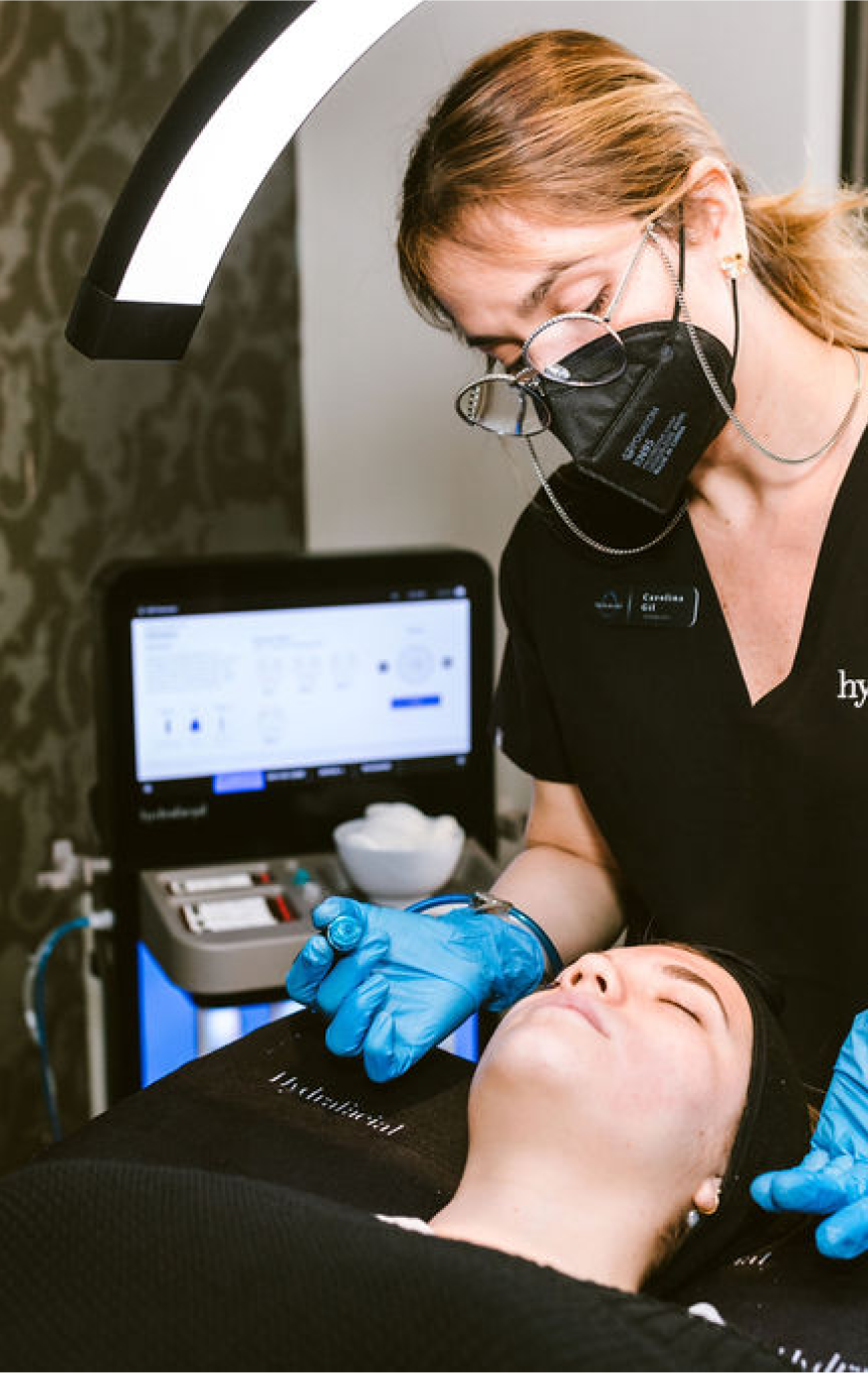 A healthcare professional wearing glasses, a black face mask, and gloves examines a patient lying down during a medical or cosmetic procedure in a clinic setting.
