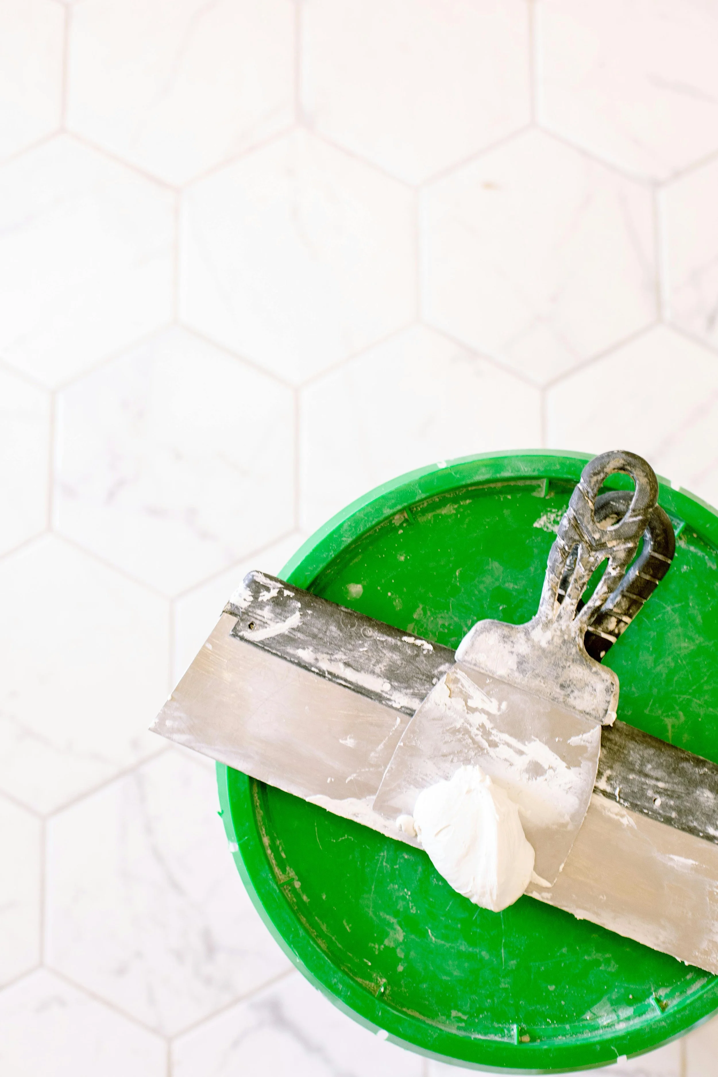 A putty knife with a dollop of white adhesive resting on a green bucket, against a white hexagonal-tile wall.