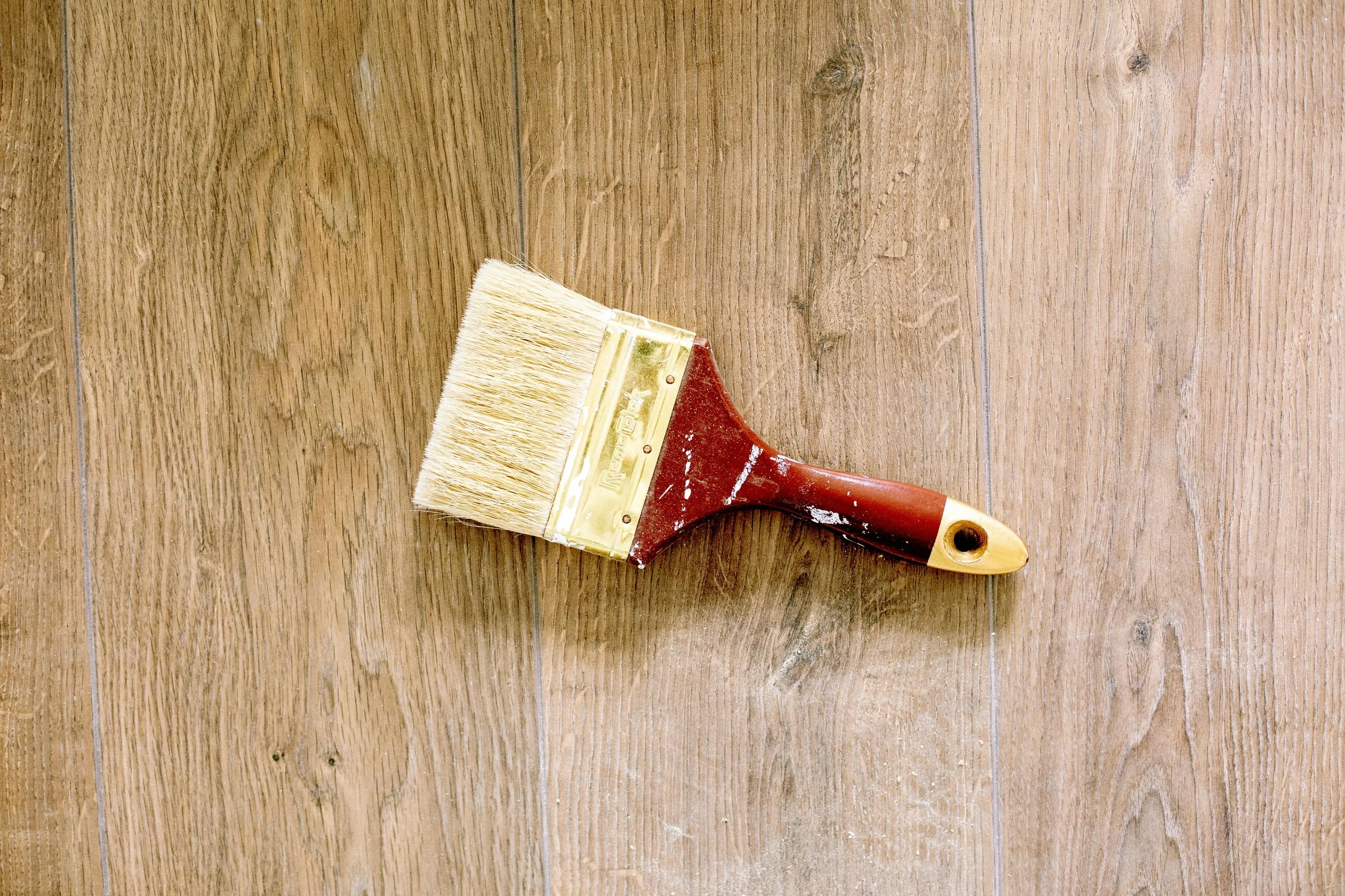 A paintbrush with a wooden handle and light-colored bristles lying on wooden flooring.