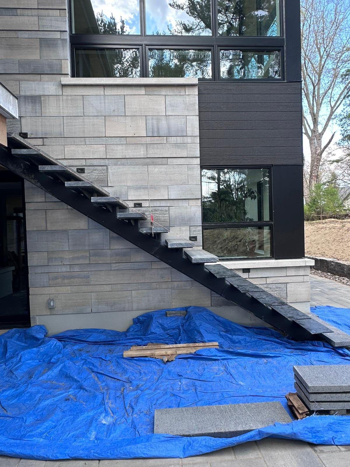 A modern building exterior under construction featuring an outdoor staircase, large windows, a gray stone wall, black wooden siding, and a blue tarp covering the ground with construction materials.