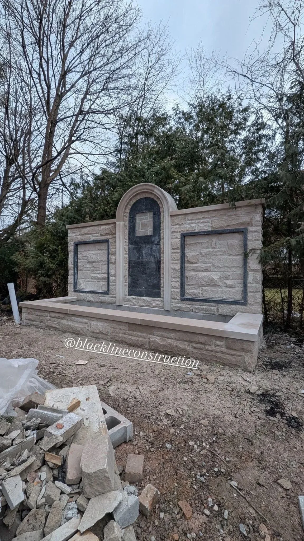 A newly constructed outdoor stone structure with a built-in shelf, set against trees and a fence, likely a decorative feature or memorial, with construction materials in the foreground.