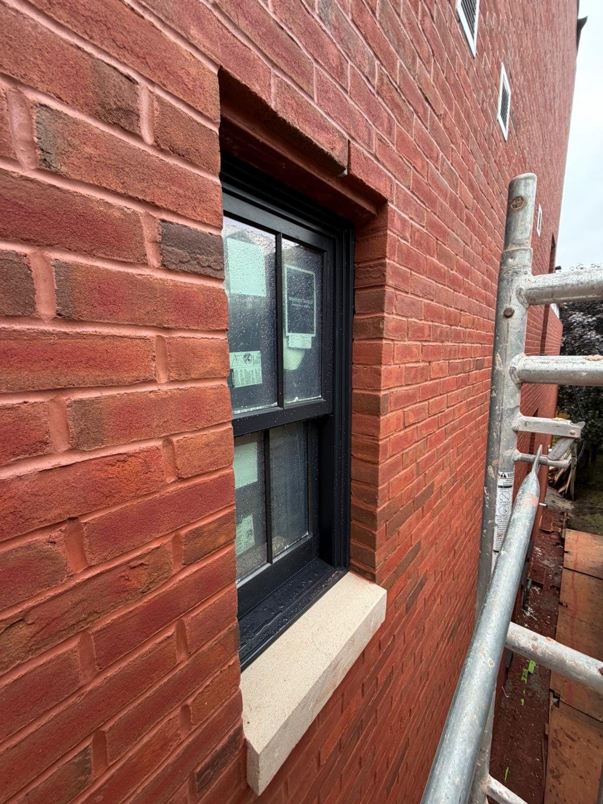 Close-up of a black-framed window set in a red brick wall, with a beige stone window sill, outside a scaffolding structure.