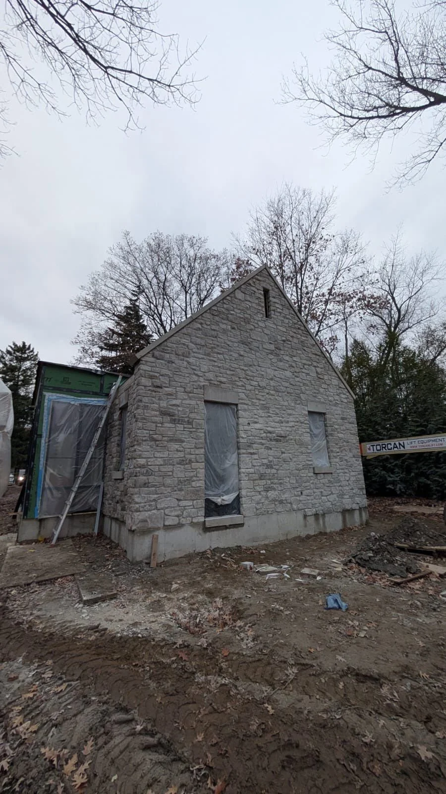 A stone house under construction with plastic-covered window openings, surrounded by a dirt ground and leafless trees against an overcast sky.