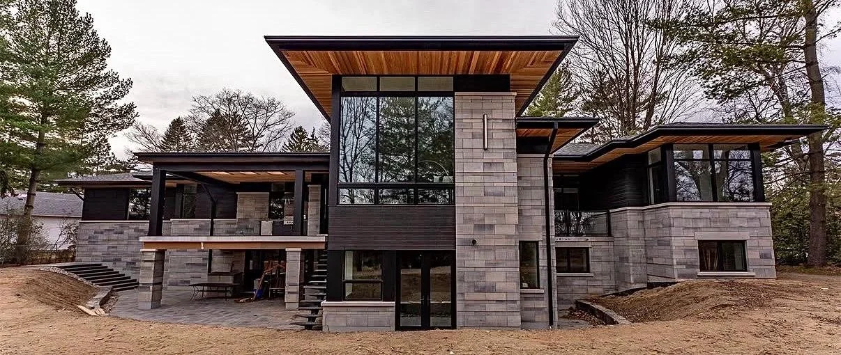 Modern two-story house with large glass windows, wooden and stone exterior, surrounded by trees.