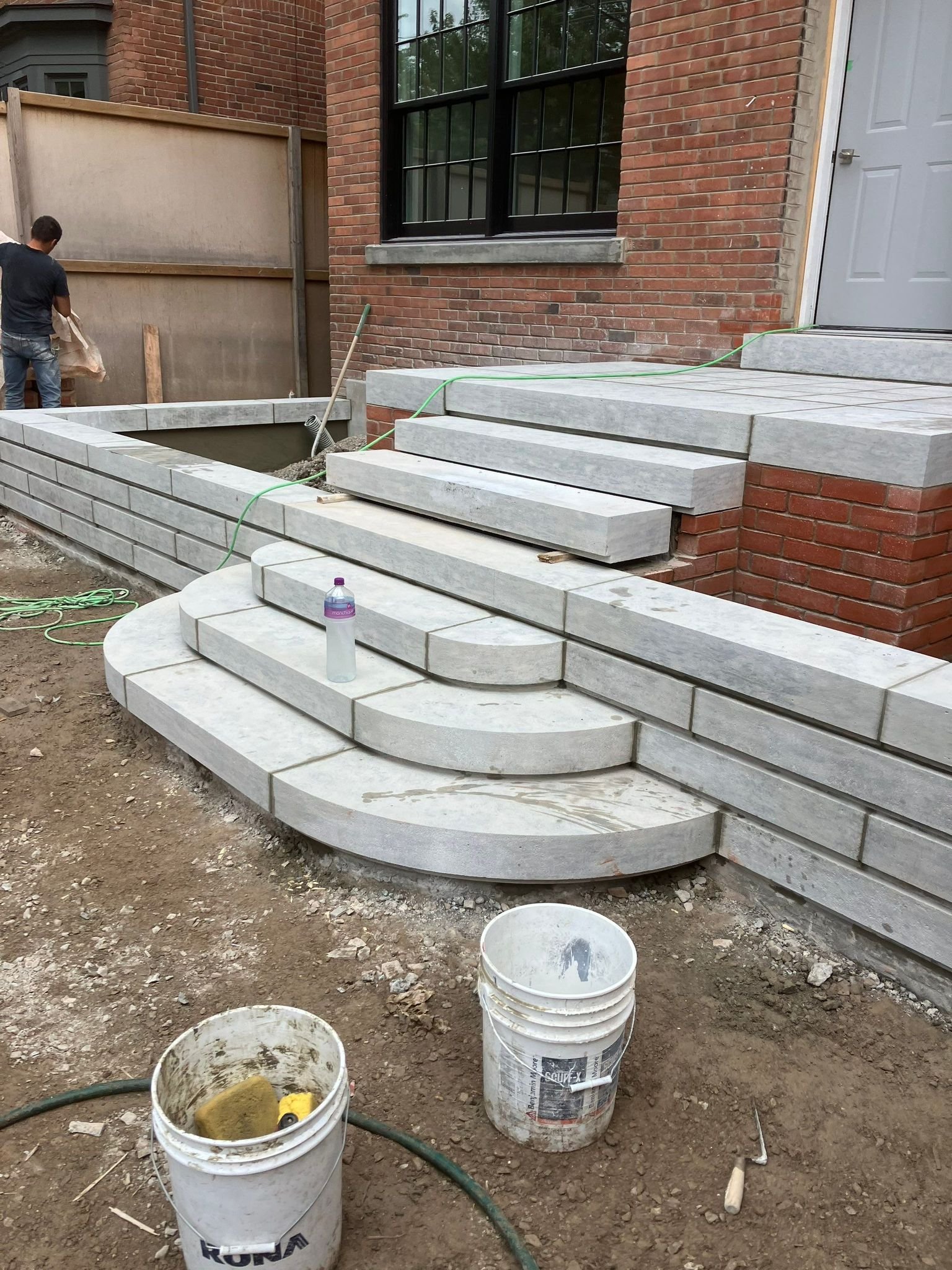 Construction site of outdoor stairs and steps with white stone tiles, with some workers and construction tools visible, in front of a brick building.