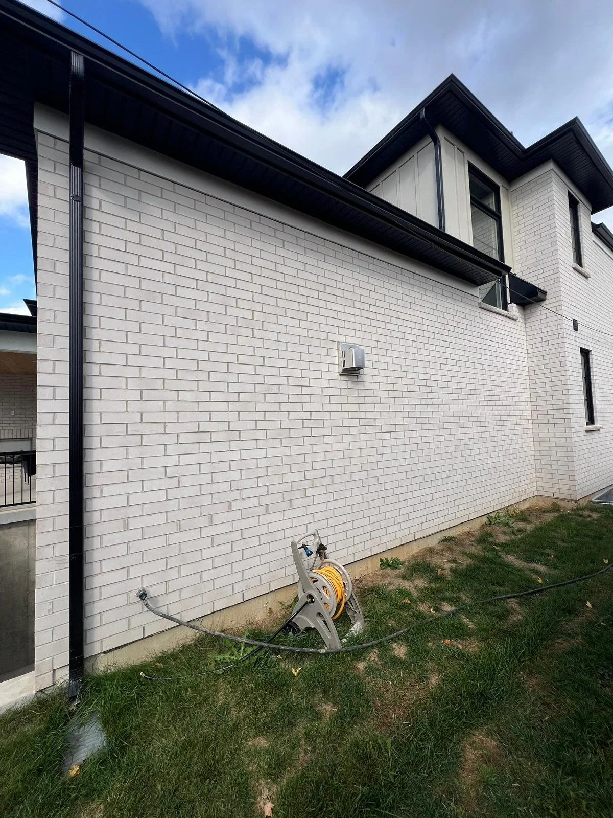 Side of a modern house with white brick exterior, black rain gutter, and a window on the upper floor. A garden hose reel is on the ground near the wall, and the sky is partly cloudy.