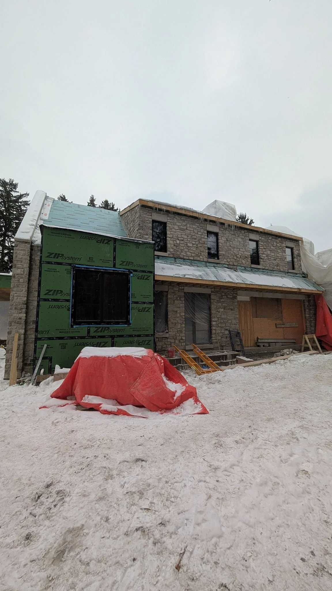 Under construction house with stone exterior, snow on ground, partially finished roof, construction materials, and tools in winter.