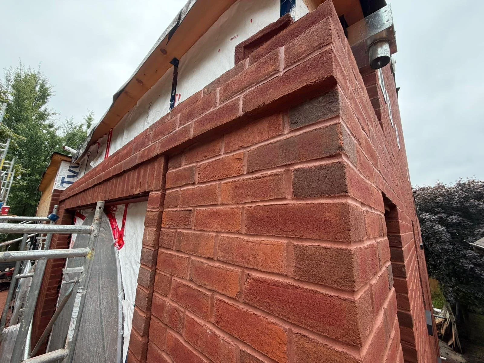 Close-up of an outdoor brick wall under construction, with scaffolding and construction materials visible nearby, and an overcast sky in the background.