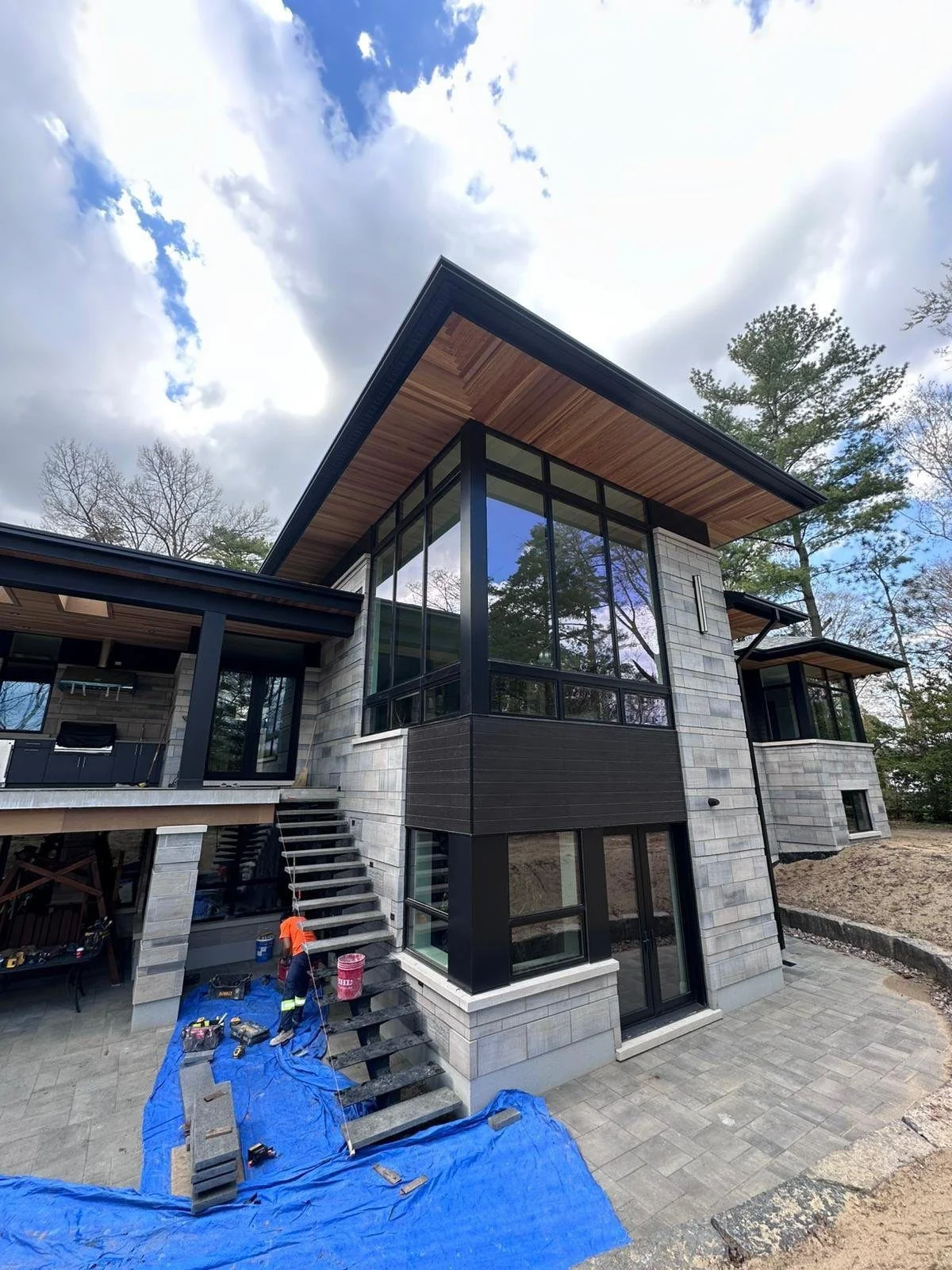 Modern house under construction with large glass windows, concrete and wood exterior, and a staircase leading to an upper door. Construction worker on site with tools and blue tarp covering the ground.