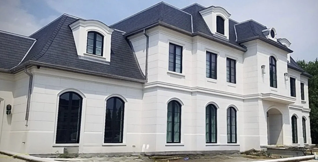 A large white mansion with multiple windows and a dark gray roof under construction.