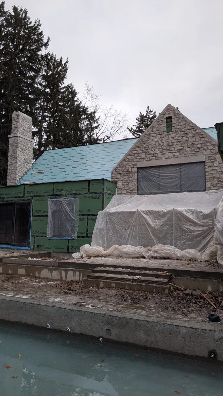 A house under construction with partially finished brick walls, a blue roof, and plastic sheeting covering windows and parts of the structure.