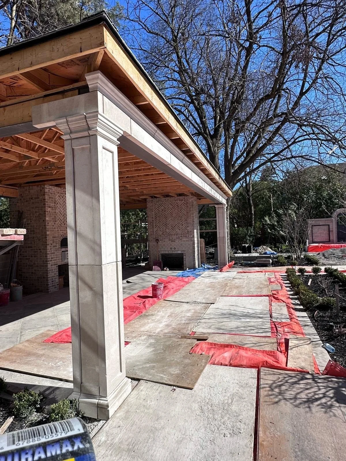 Construction site of an outdoor structure with brick columns, wooden framework, and a stone patio, during daytime with leafless trees and blue sky in the background.