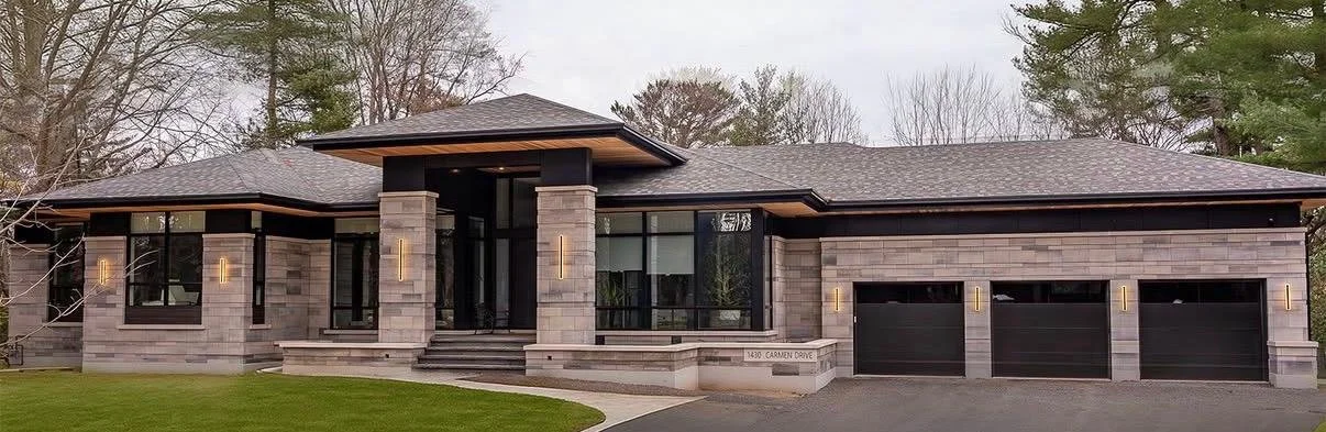 Modern single-story house with a dark roof and gray brick exterior, featuring large windows, three garage doors, and a well-maintained lawn.