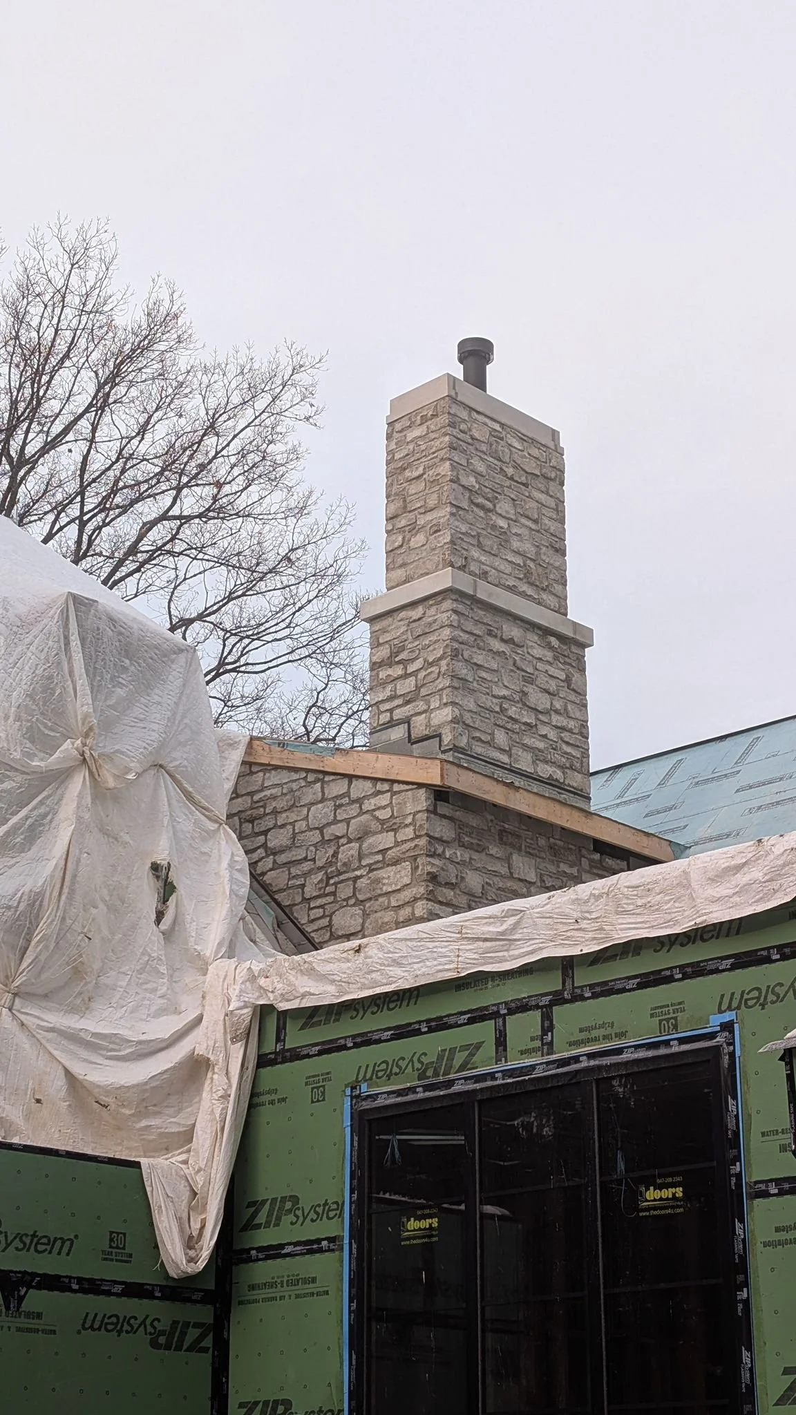Construction site with a finished brick chimney and a partly covered building with green insulation wrap and a black window.