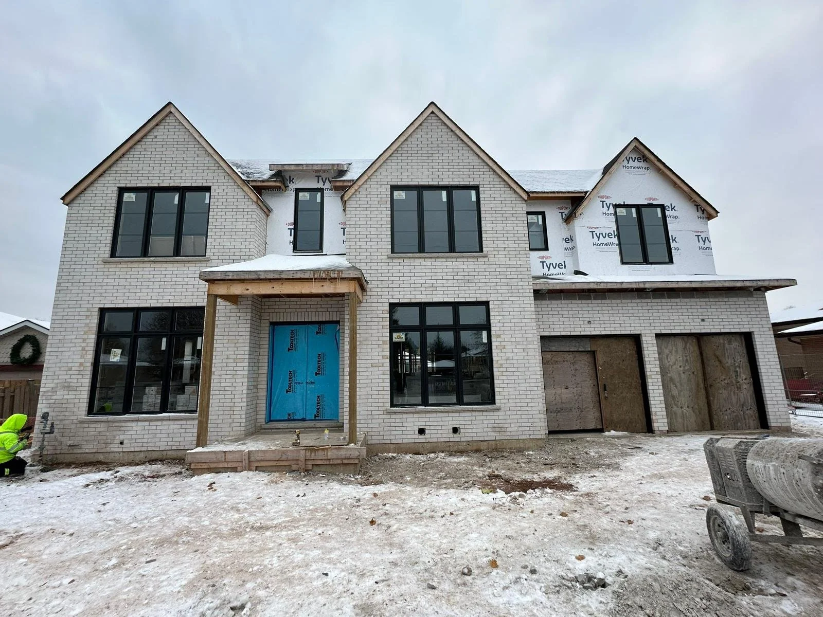 A large two-story house under construction with a snow-covered yard, unfinished garage, and scaffolding around the front door.