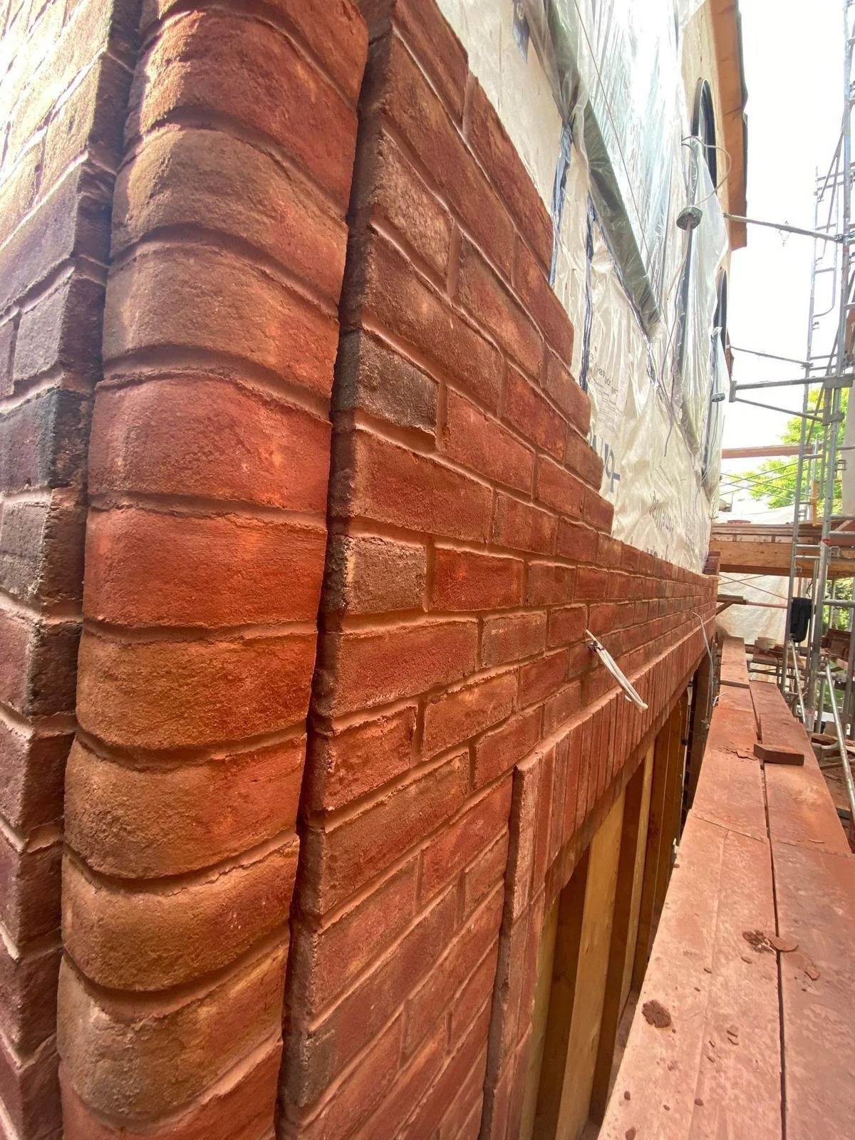 Close-up of a brick wall under construction, showing a detailed corner with bricks and mortar, with scaffolding and building wrap visible in the background.
