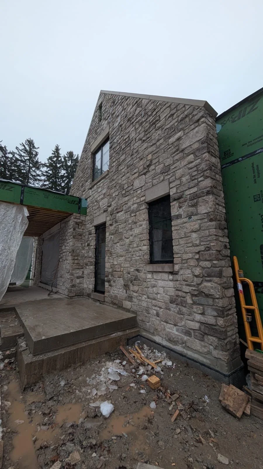 View of a stone residential building under construction with three visible windows, a small concrete porch, and construction materials and debris around the site.