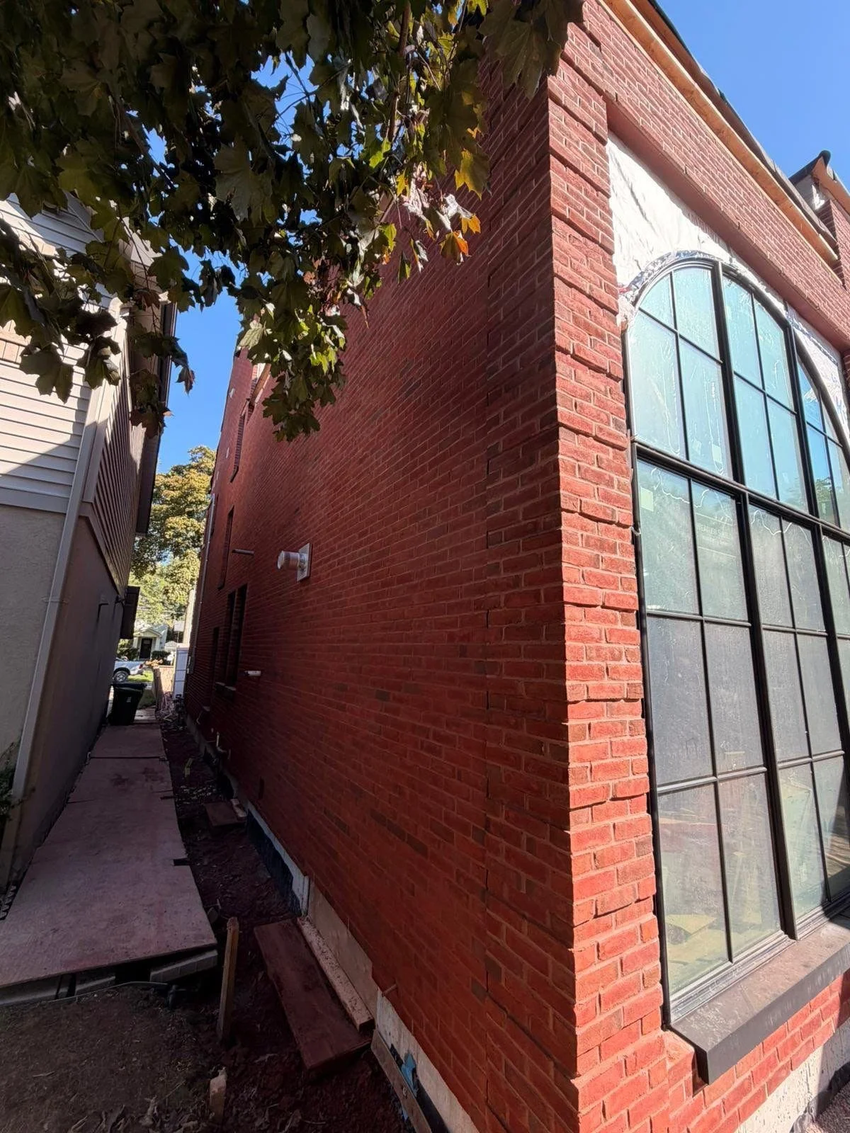 Exterior view of a brick building with large arched windows on the right, a tree in the upper left corner, and a narrow alleyway with construction materials on the left side, under a clear blue sky.
