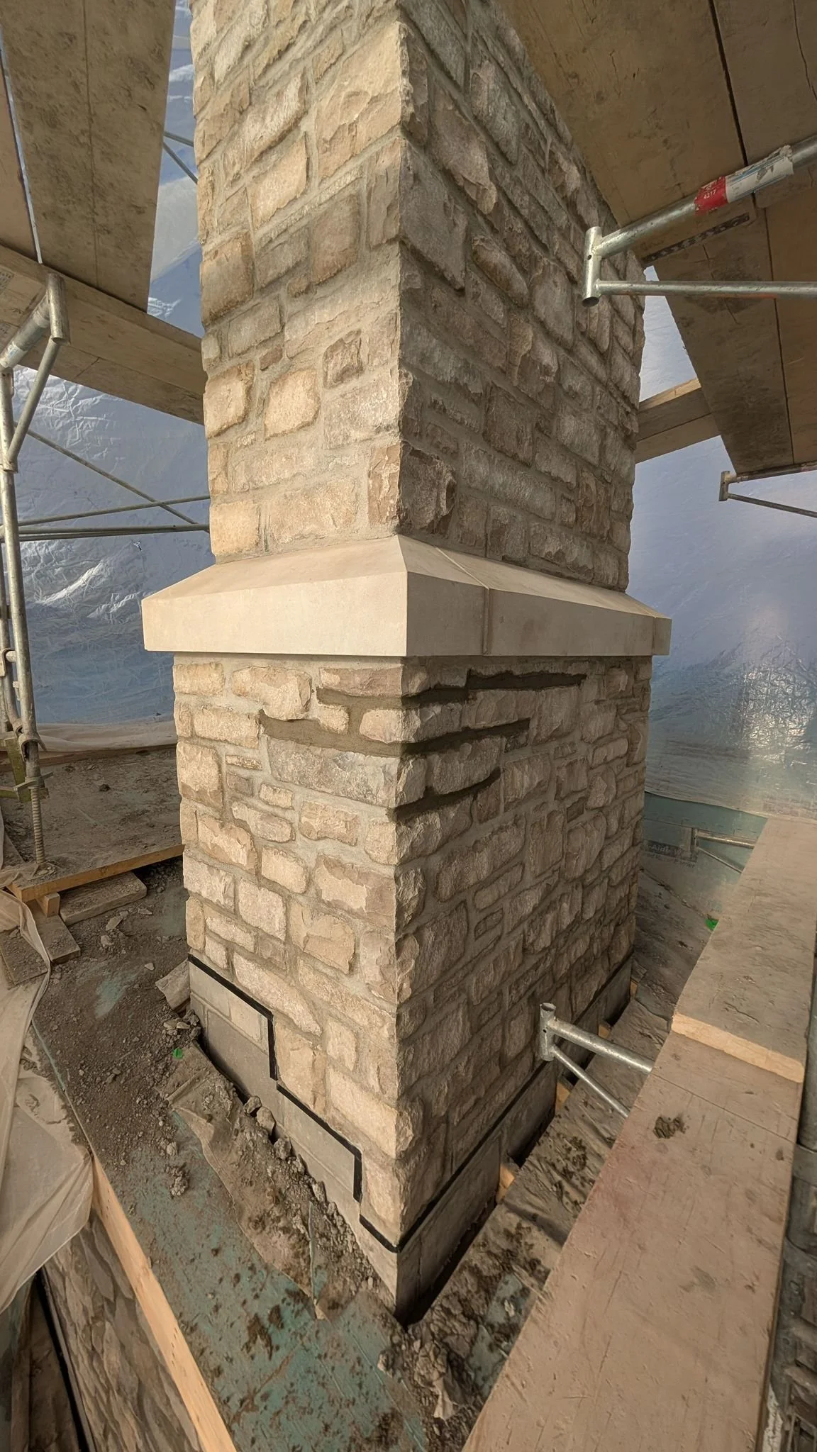 A brick chimney with a stone cap on a construction site, surrounded by scaffolding and construction materials.