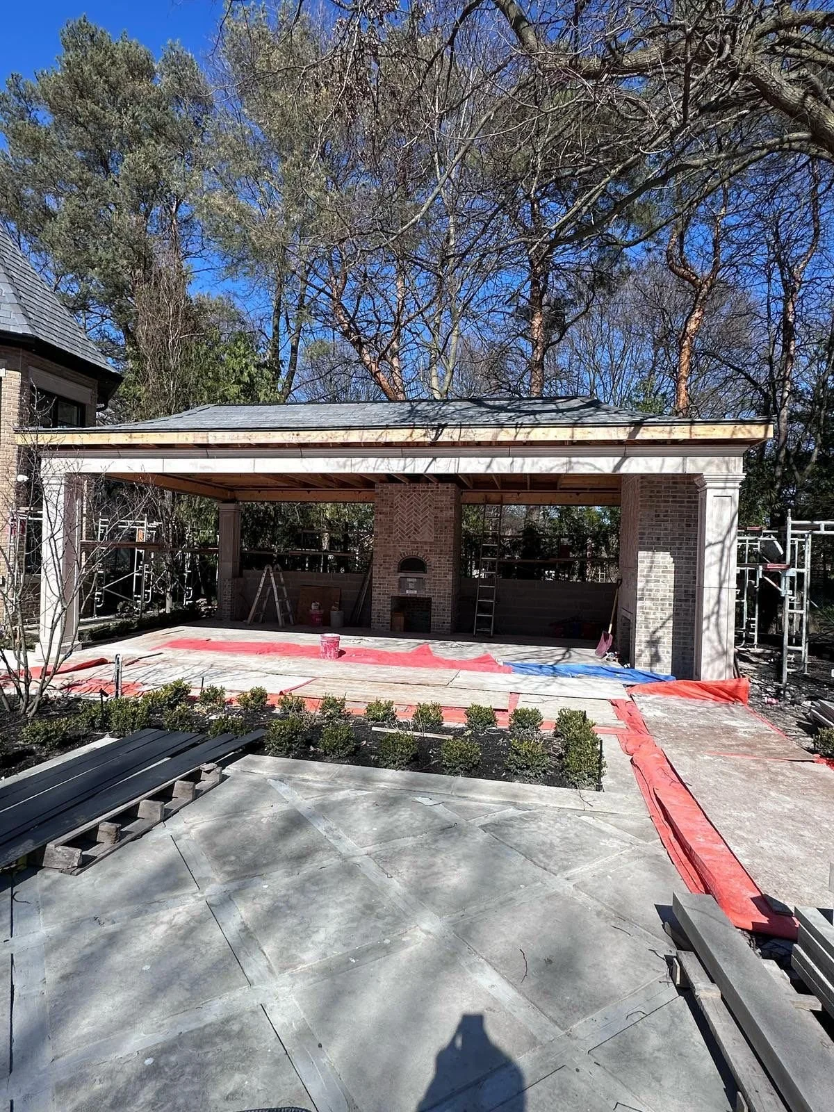 Under construction outdoor patio with brick fireplace, covered with a roof, surrounded by trees, and various building materials and tools nearby.