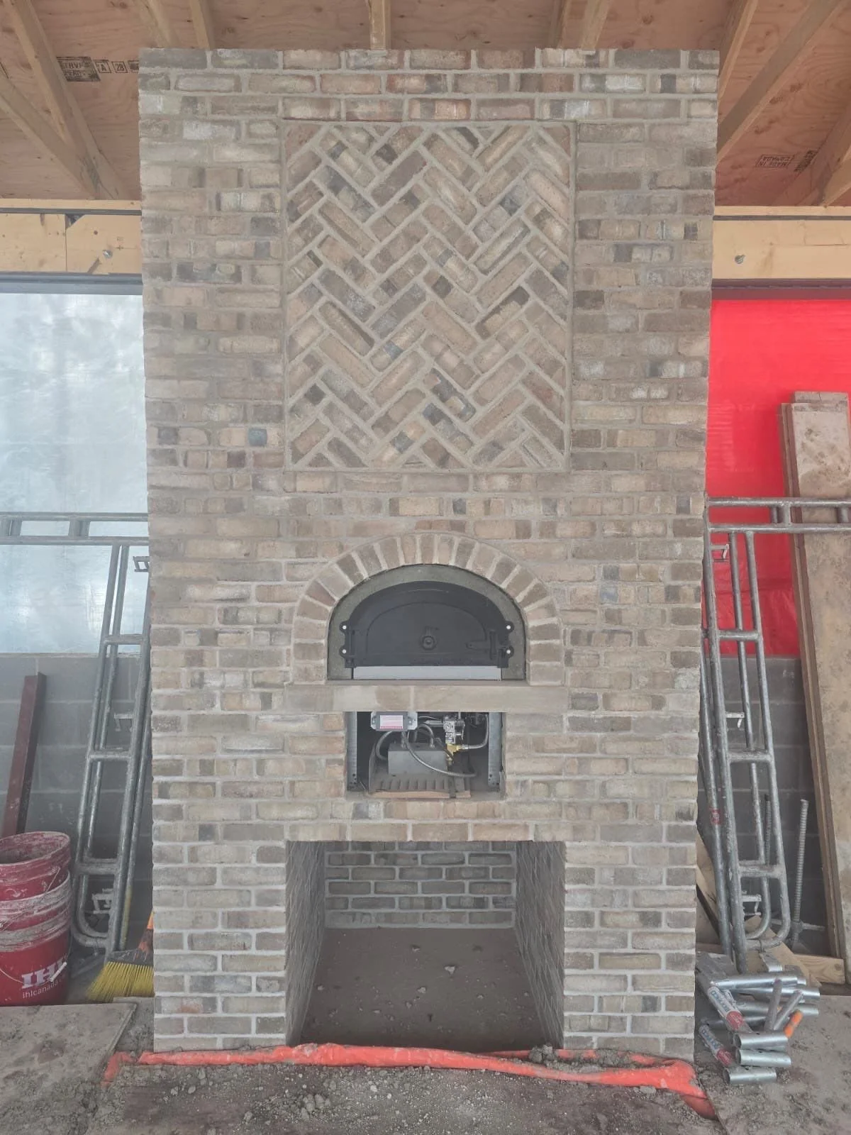 Under construction brick fireplace with a chimney, partially installed oven door, and construction materials around.