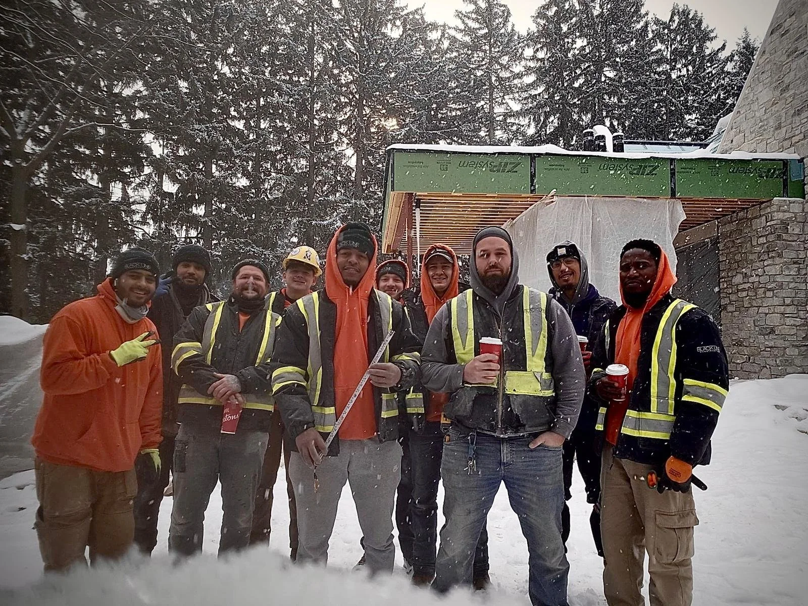 Group of construction workers standing in snow outside, holding beverages, wearing safety gear, and smiling.
