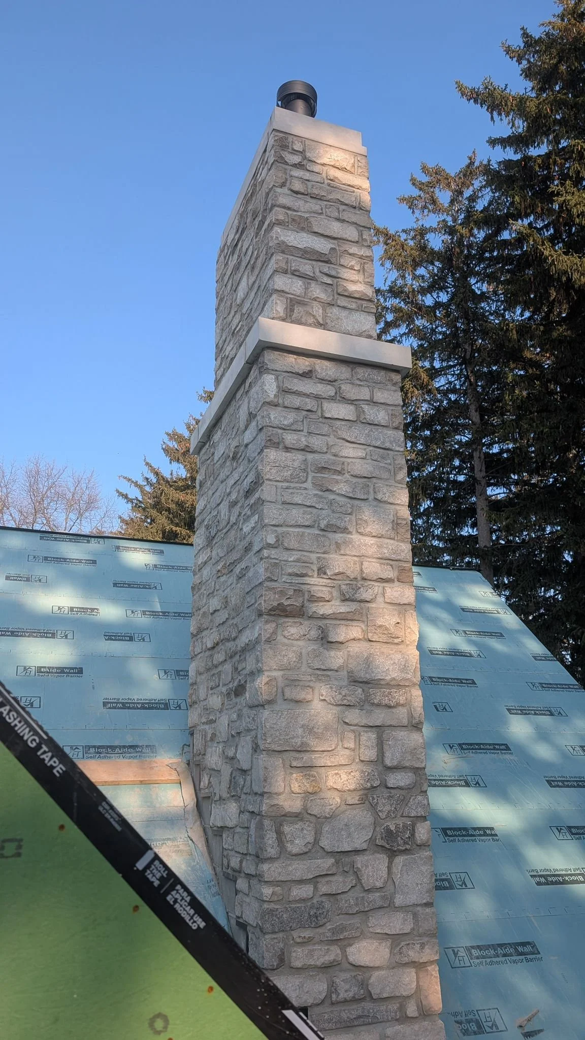A large stone chimney with a cap, standing beside the roof of a house, during a clear day with a blue sky and trees in the background.