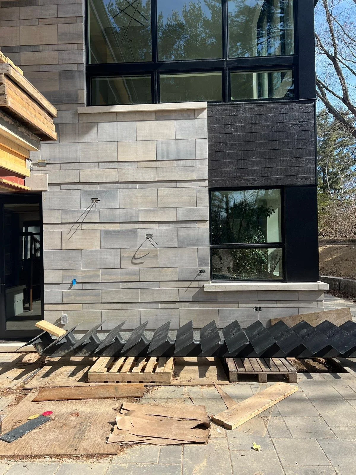 Building exterior under construction, featuring a modern design with partly finished gray and black stone walls, large windows, and construction materials and tools on the ground.