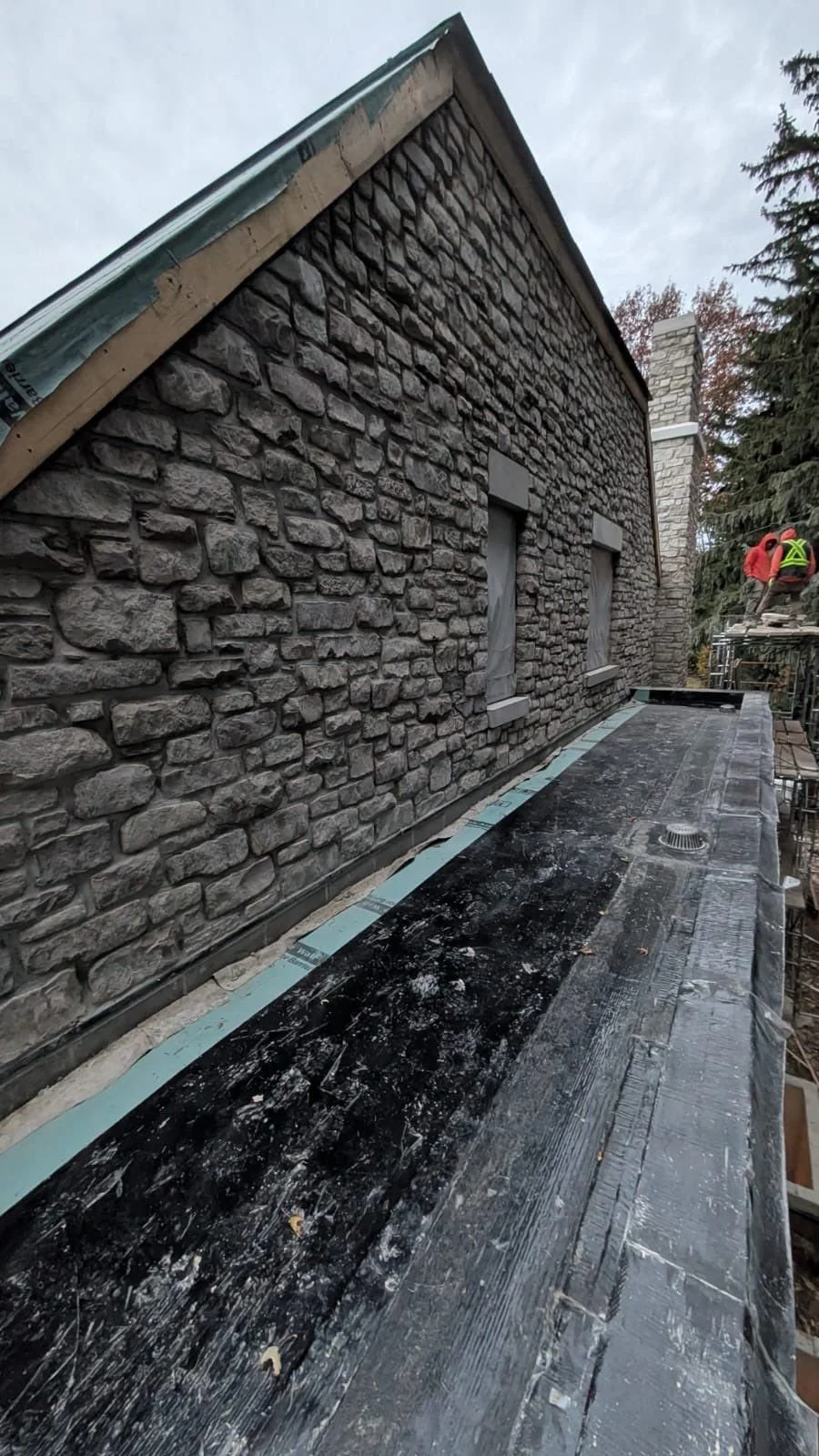 Construction workers on scaffolding working on a stone exterior wall of a building with three recently installed windows, and a roof edge with green trim.