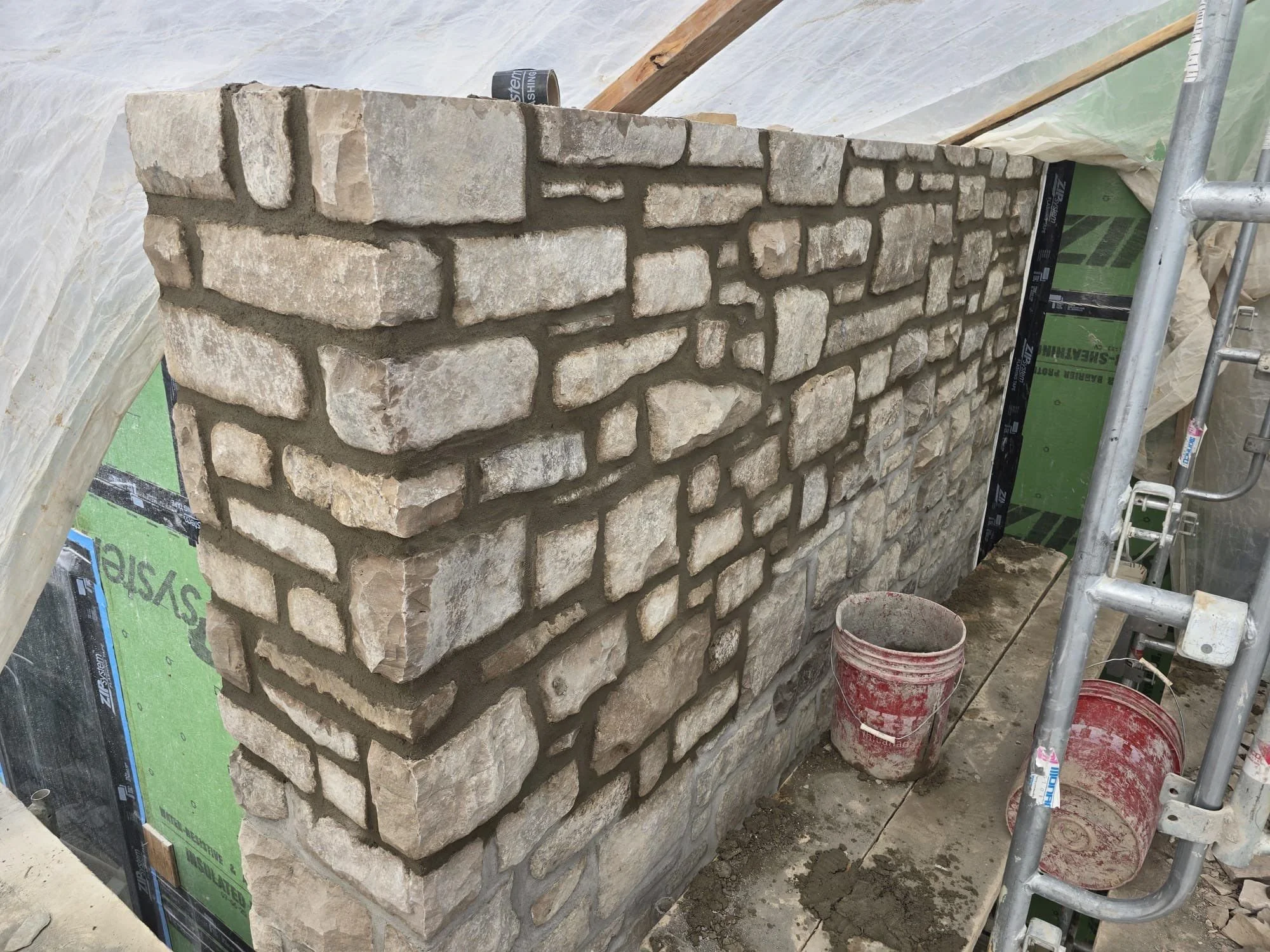 A stone wall under construction with beige and light brown stones, surrounded by construction scaffolding and tools, including buckets and plastic sheeting.