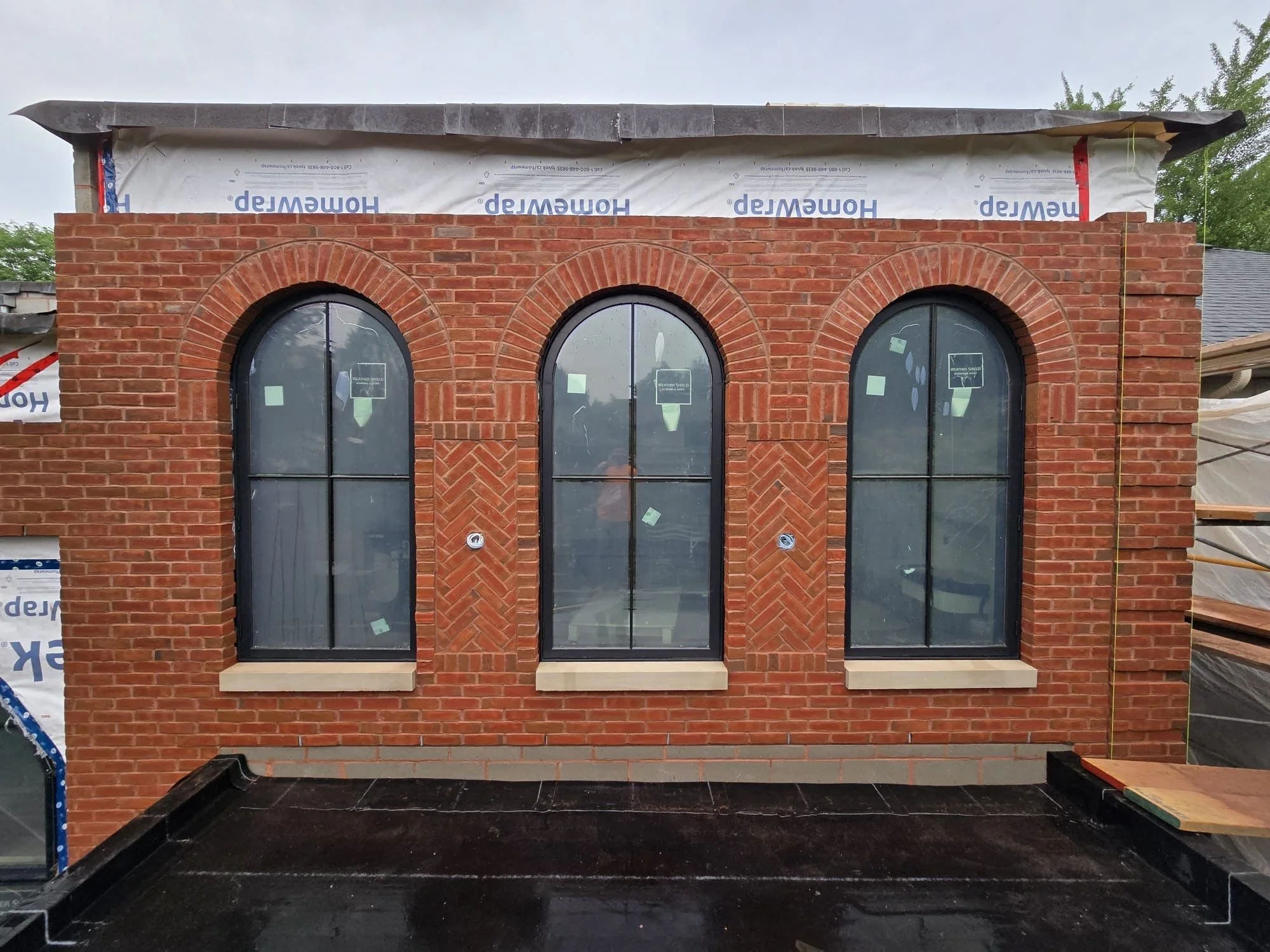 Brick building under construction with three tall arched windows and black frames, with some construction materials and insulation visible at the top and sides.